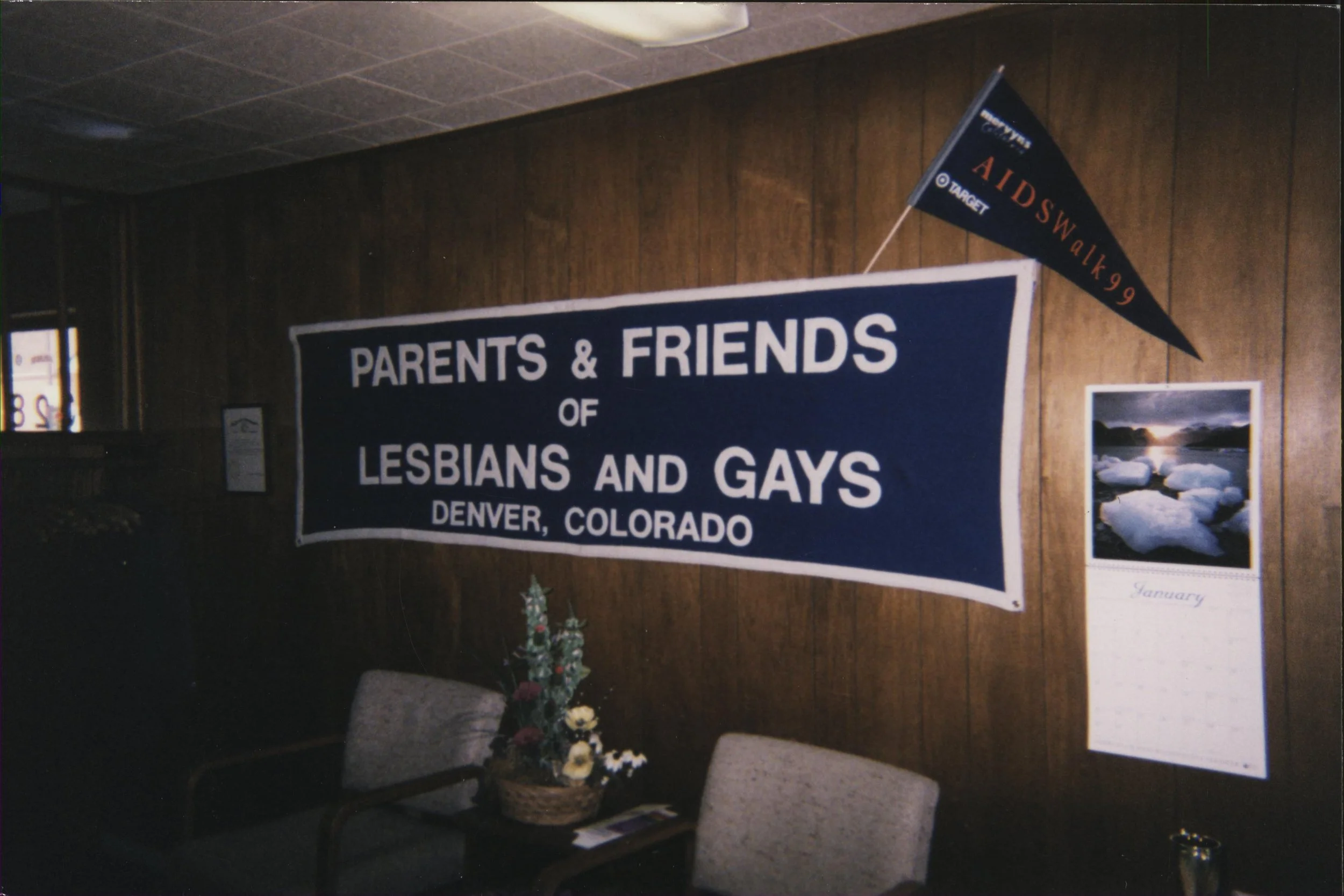 Banner reading 'Parents & Friends of Lesbians and Gays Denver, Colorado' on a wooden wall with a calendar and a small flag that says 'AIDS Walk 99' and a calendar displaying January.