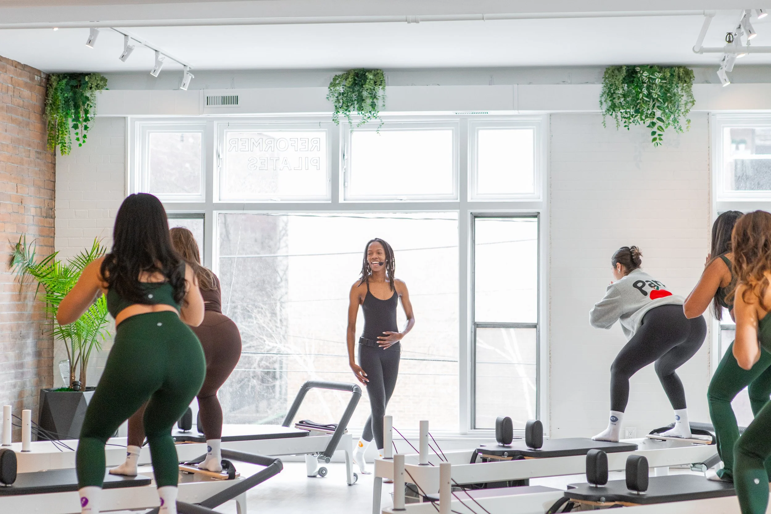 Women participating in a Pilates class led by an instructor in a bright studio with large windows, white walls, and green plants.