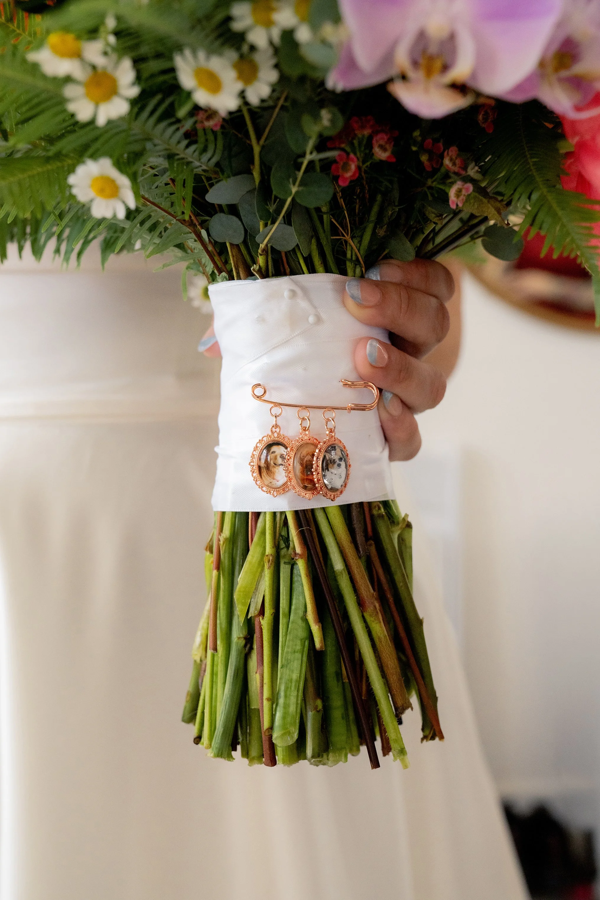 close up detailed image of the bottom portion of a wedding bouquet to focus on the pin at the base that showcases the brides three dogs in individual gold frames
