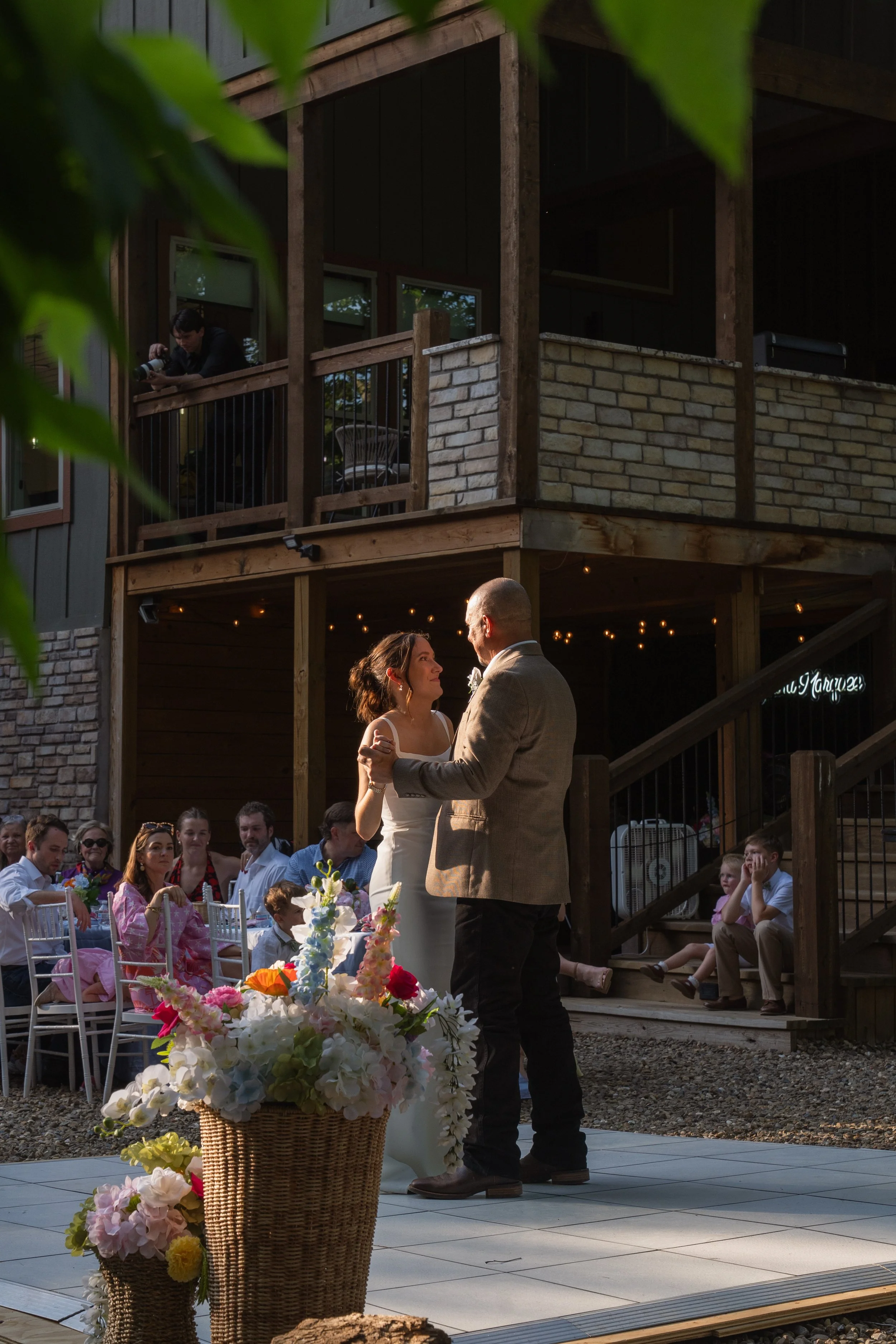 image of a bride and her father dancing together on an otherwise empty stage as family and friends look from beyond with a cabin and foliage in the background and golden sunlight shining upon the both of them and the bride is smiling