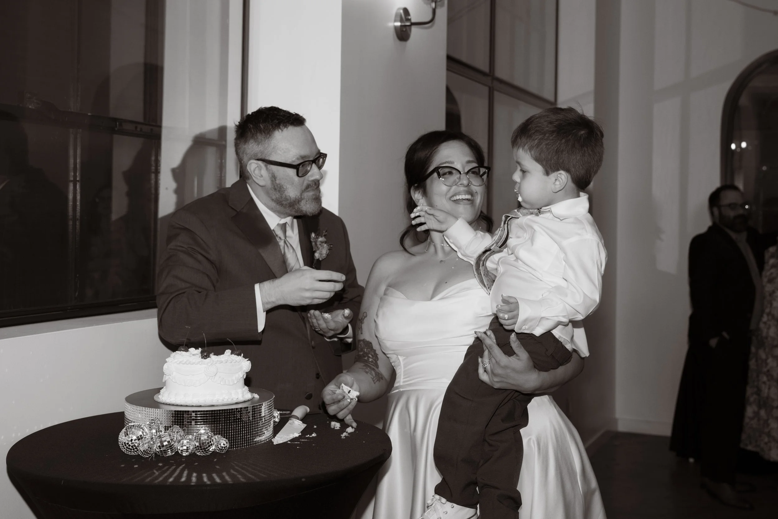 black and white image of a couple dressed in wedding attire with a suit and white wedding dress in front of their wedding cake, holding their toddler sun as they give him a piece of the cake, all are smiling