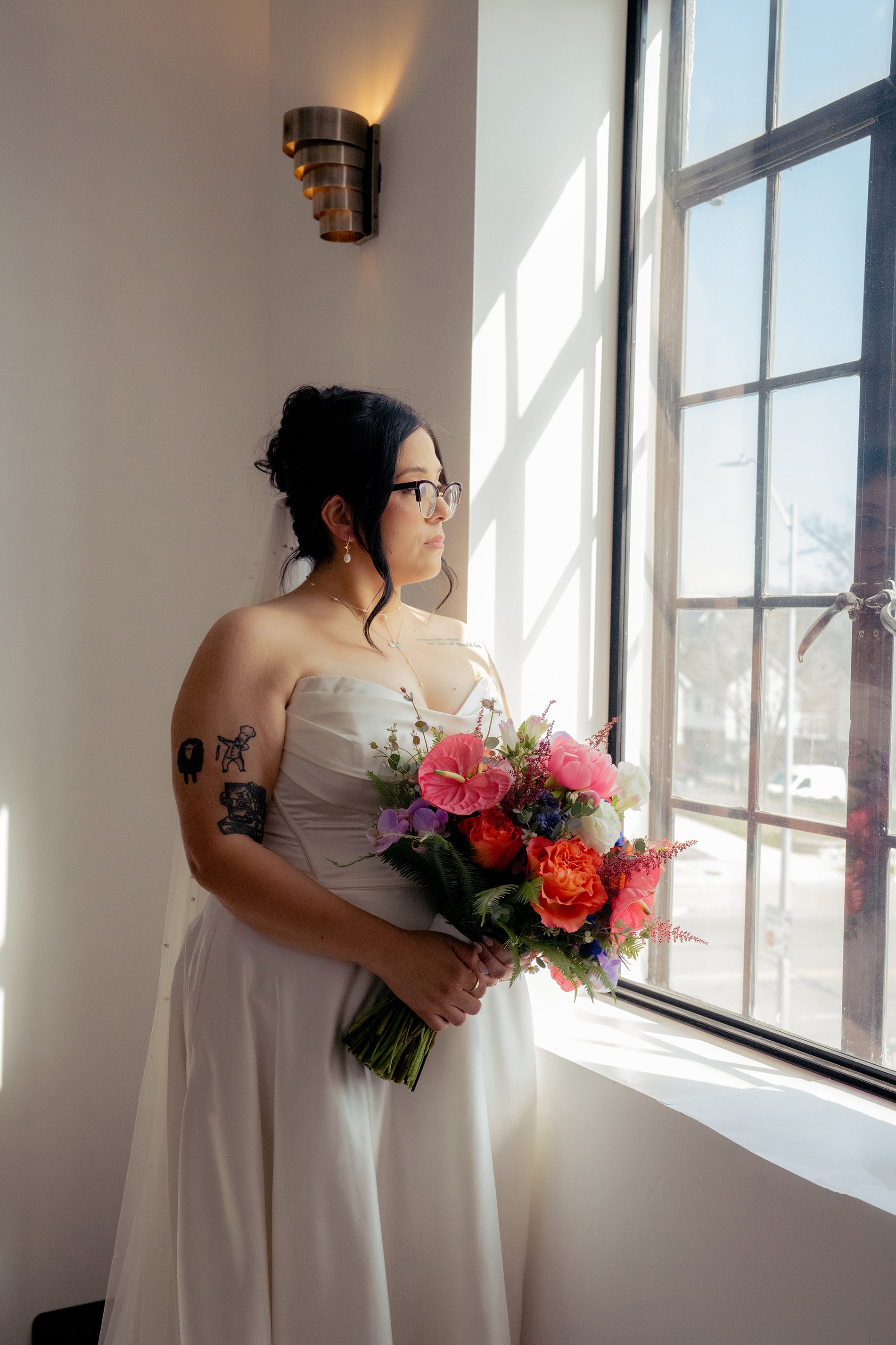 woman in a white dress holding a bouquet of prink and orange flowers as she stares out the window, basking in soft lighting from the sun shining in outside, film inspired editing with yellow and rose hues