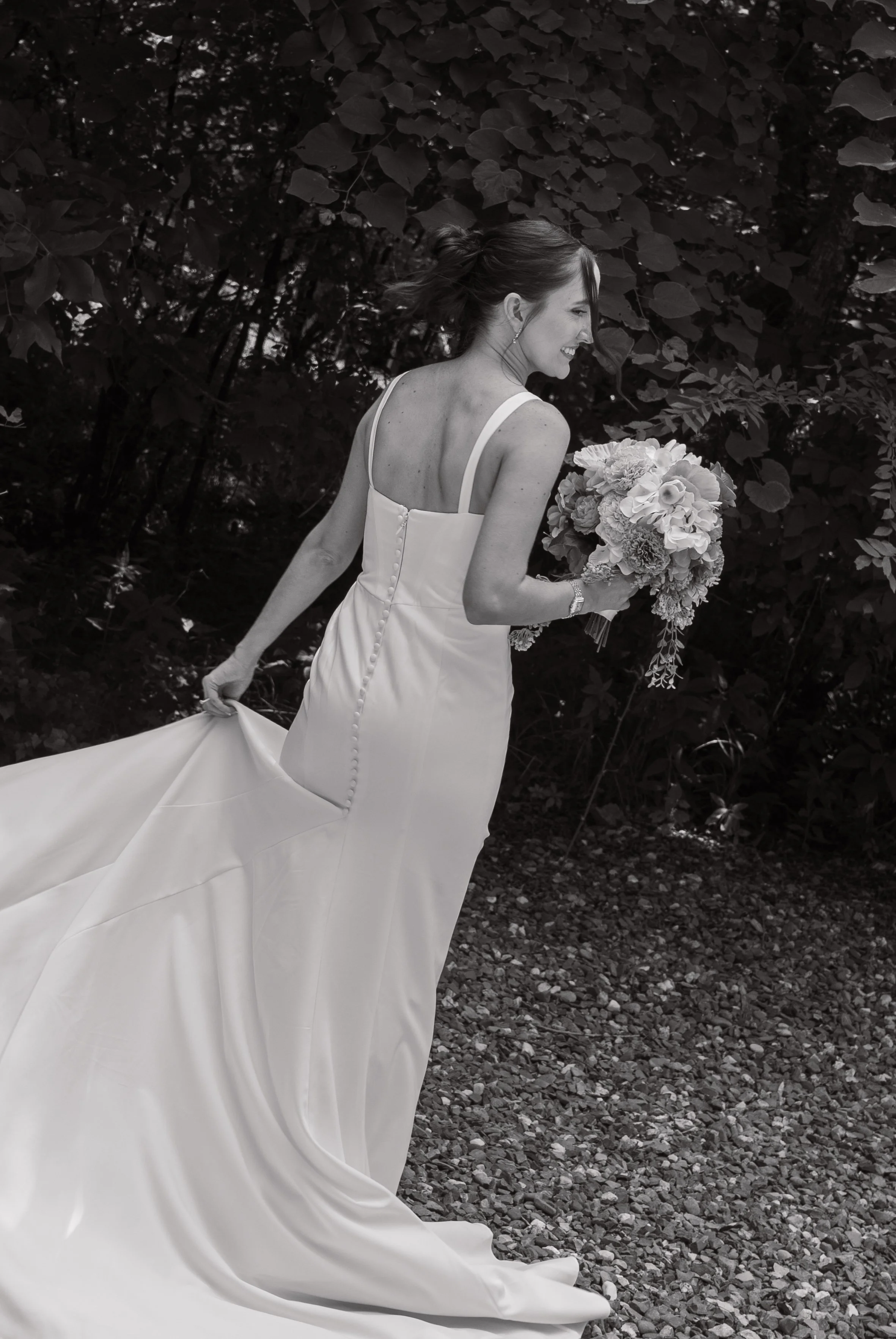 black and white image of bride in an elegant white dress spinning, train flowing behind as the details of the buttons along the back of her dress show, she's peeking over her shoulder smiling and holding her bouquet in hand