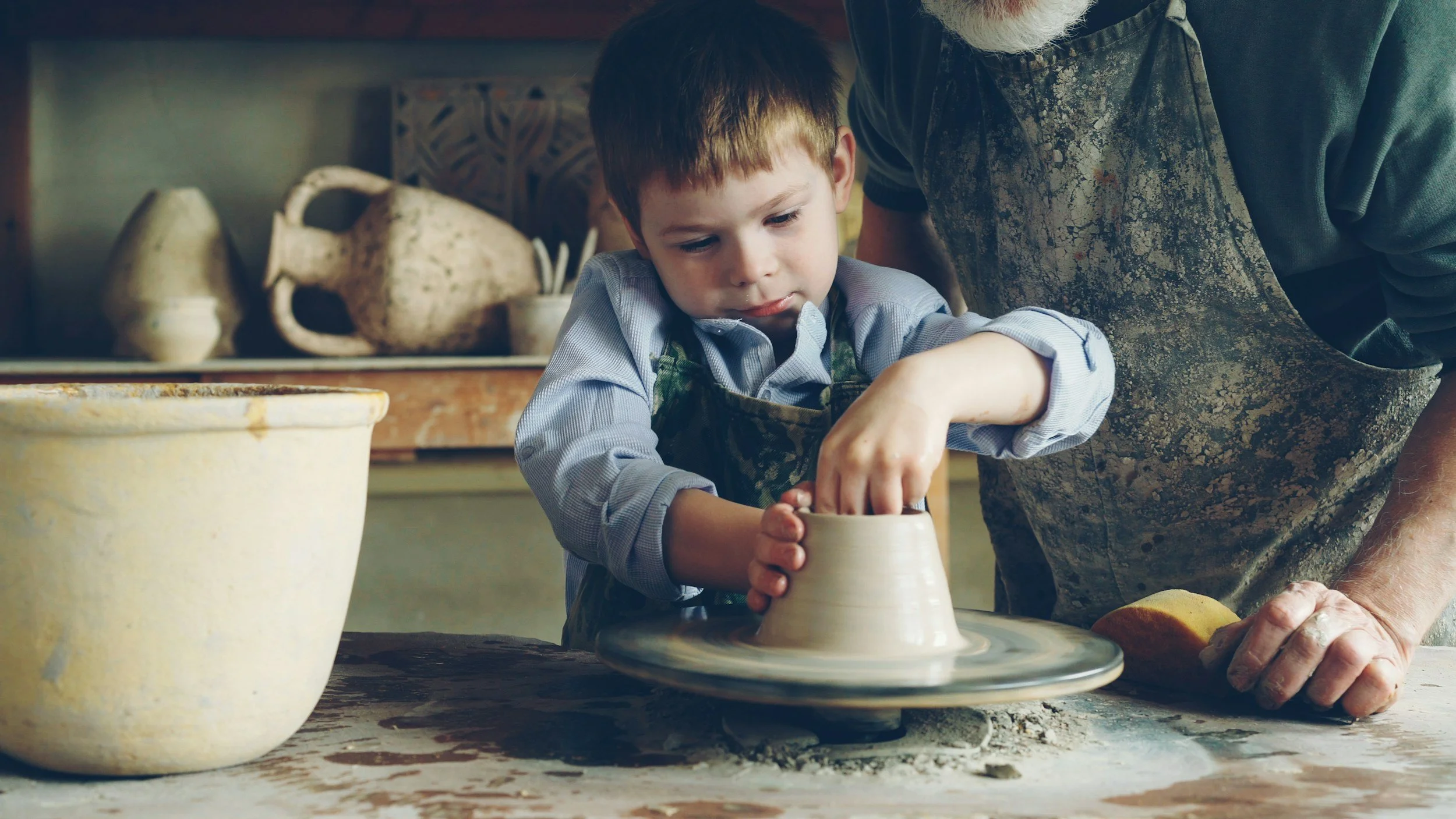 A young boy and an older man working together on a pottery wheel to shape a clay pot in a ceramics studio.