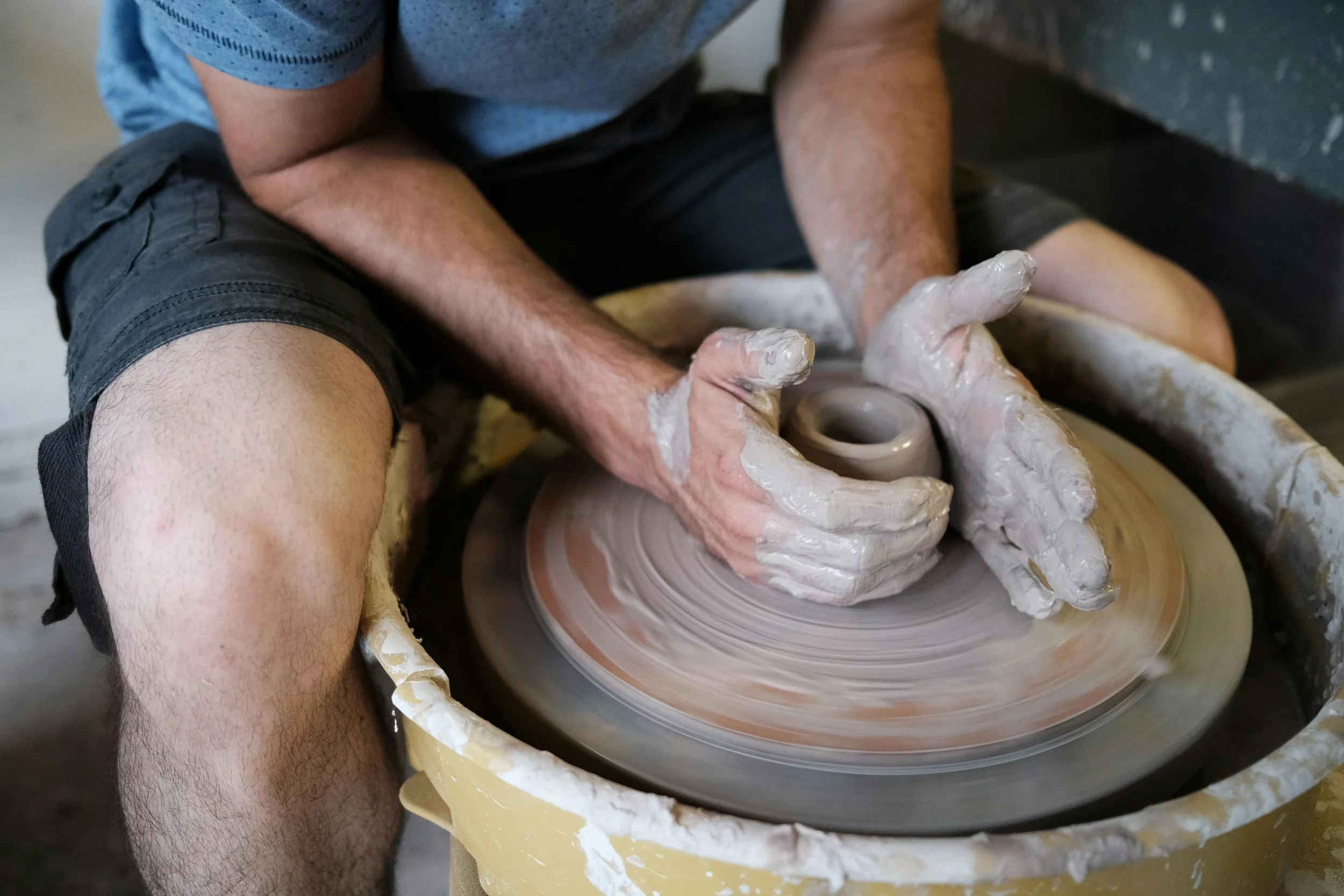 A person shaping clay on a pottery wheel, with hands covered in clay, wearing a grey shirt and black shorts.