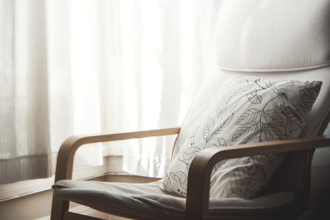 Minimalist interior scene featuring a wooden armchair with a patterned cushion beside sheer curtains, softly lit by natural light for a calm and restful atmosphere.