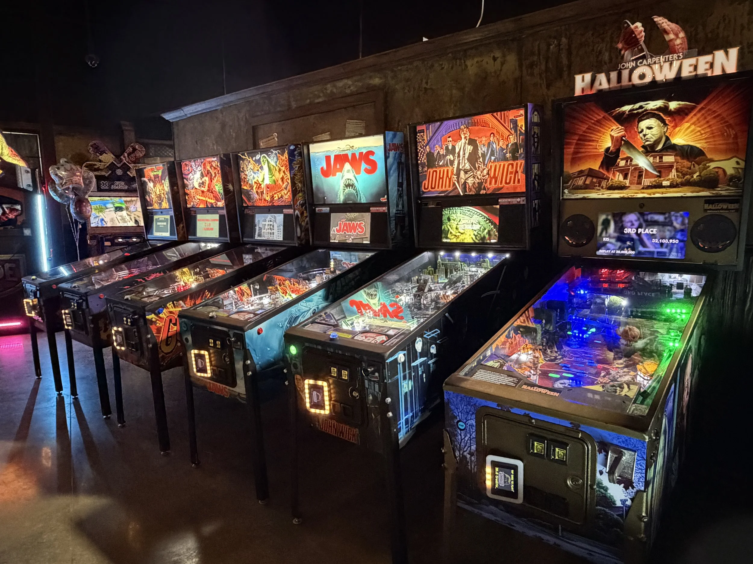 six pinball machines lined up against a wall at an arcade with neon lights on their signs