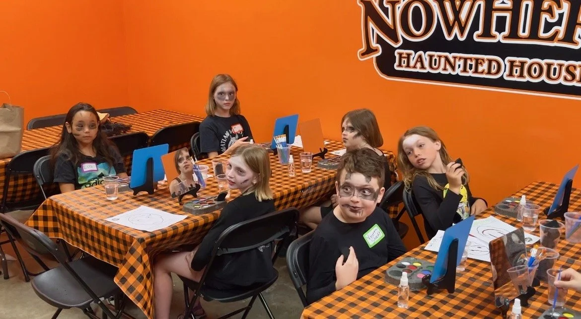 Children with face paint sitting at tables with Halloween-themed decorations, participating in a youth haunt actor activity with makeup supplies and mirrors, in front of an orange wall with Halloween signage.