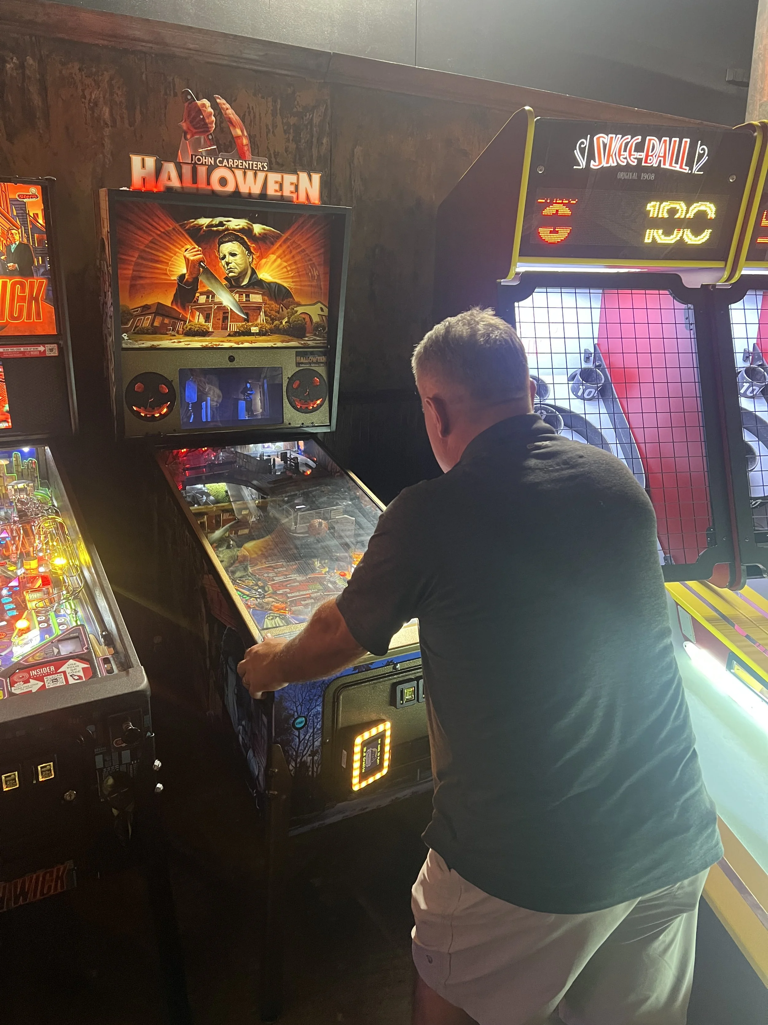 A man playing a Halloween pinball machine in an arcade with other pinball machines around him 