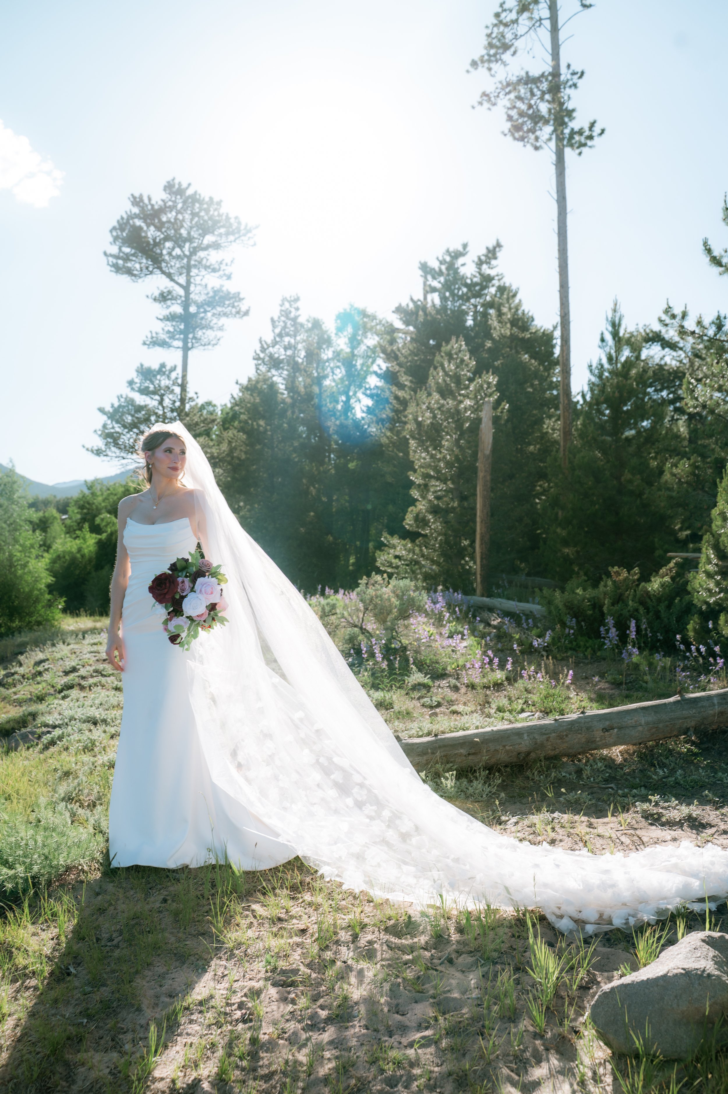 A bride in a white wedding dress holding a bouquet of roses and greenery, standing outdoors in a natural setting with trees and purple flowers, under bright sunlight.