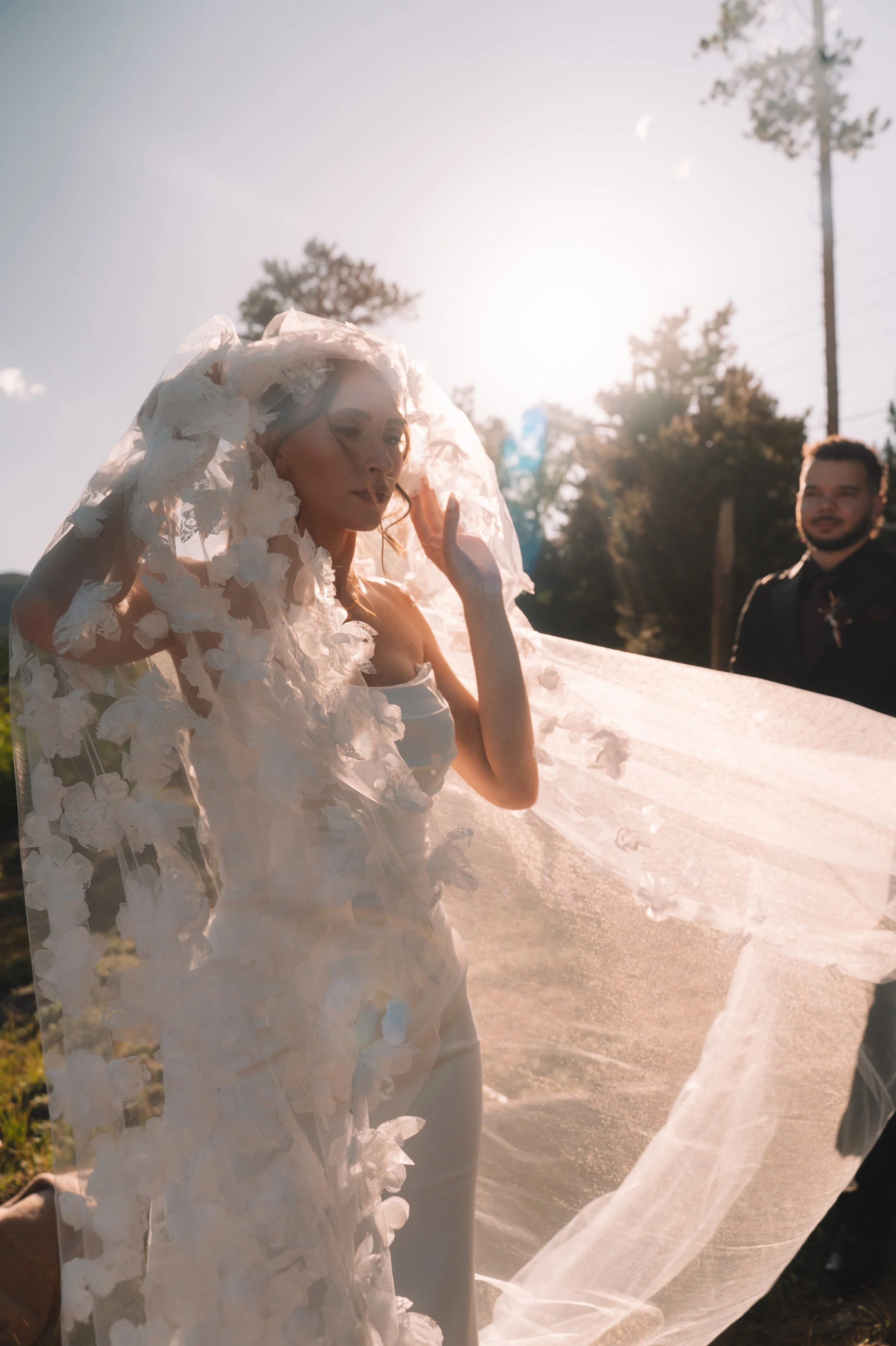 A woman in a white dress with floral details, wearing a matching veil, stands outdoors with sunlight shining behind her. A man in dark clothing is visible in the background, along with trees and a clear sky.