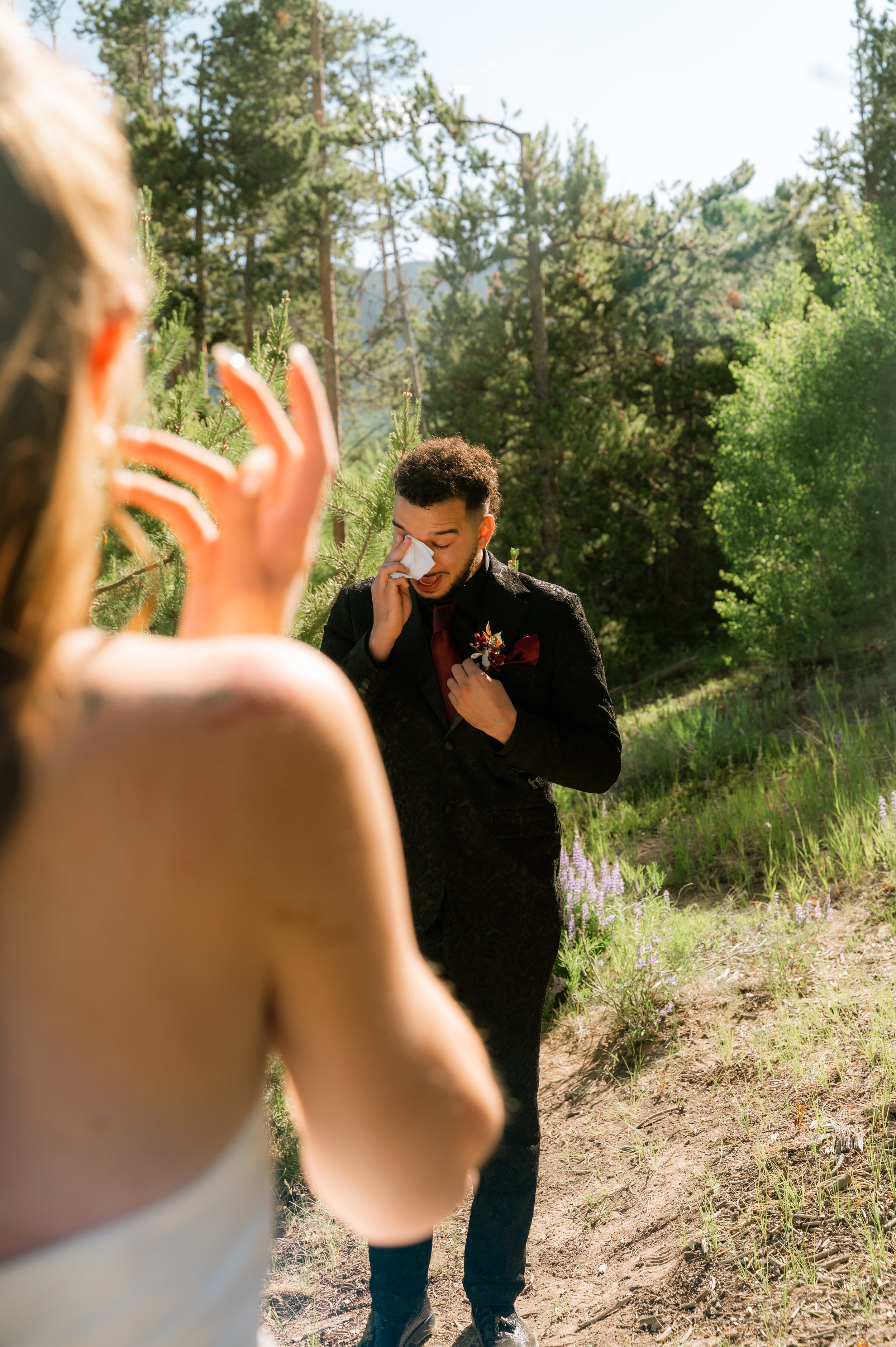 A man dressed in a black suit with a maroon tie and boutonniere, standing outdoors in a forested area, wiping his eye with a tissue, likely emotional, with a blurred figure in the foreground holding up a hand.