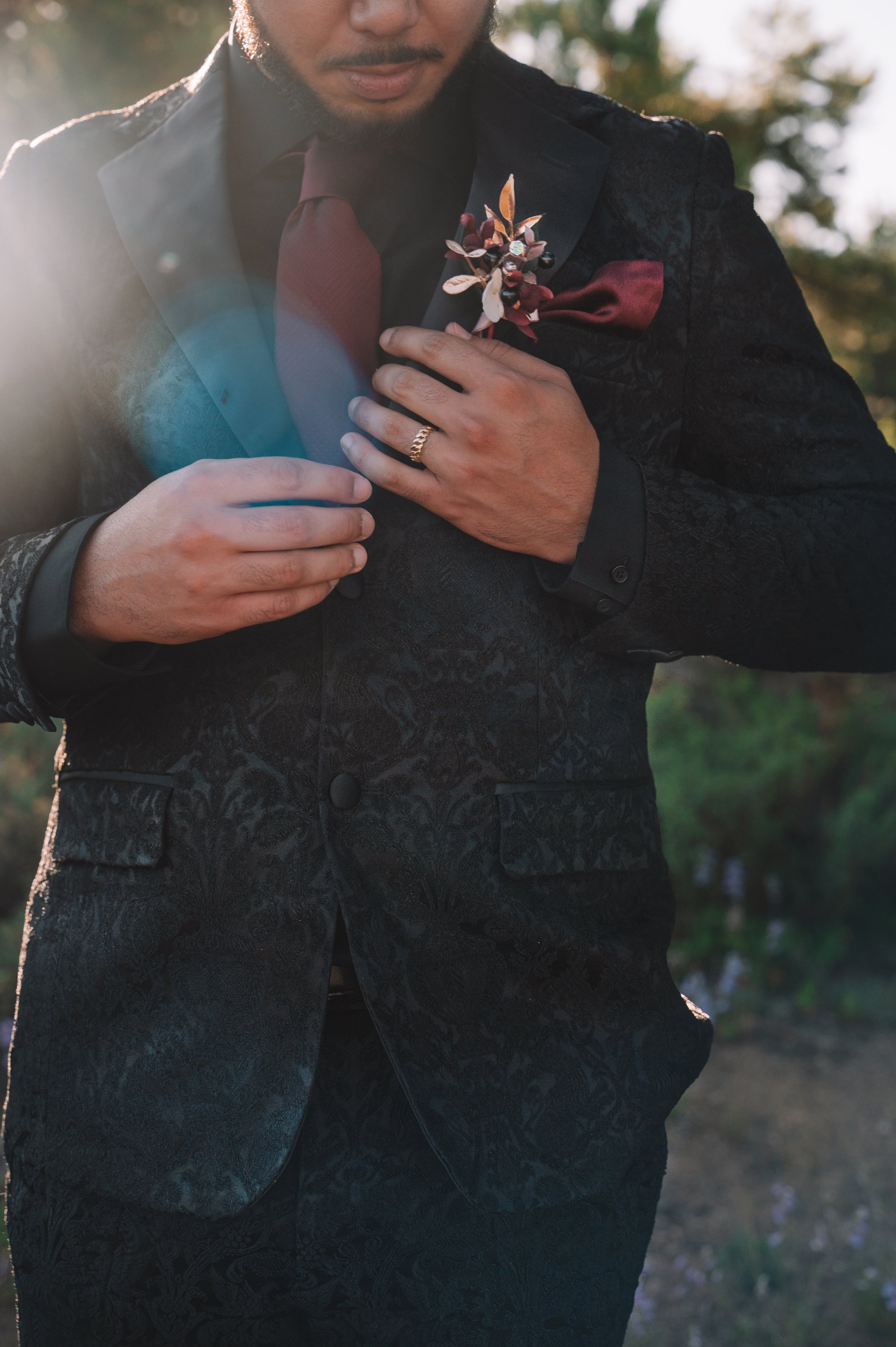 A man adjusting his black patterned suit jacket outdoors, wearing a maroon shirt, matching maroon tie, and a boutonnière on his lapel, with sunlight and trees in the background.