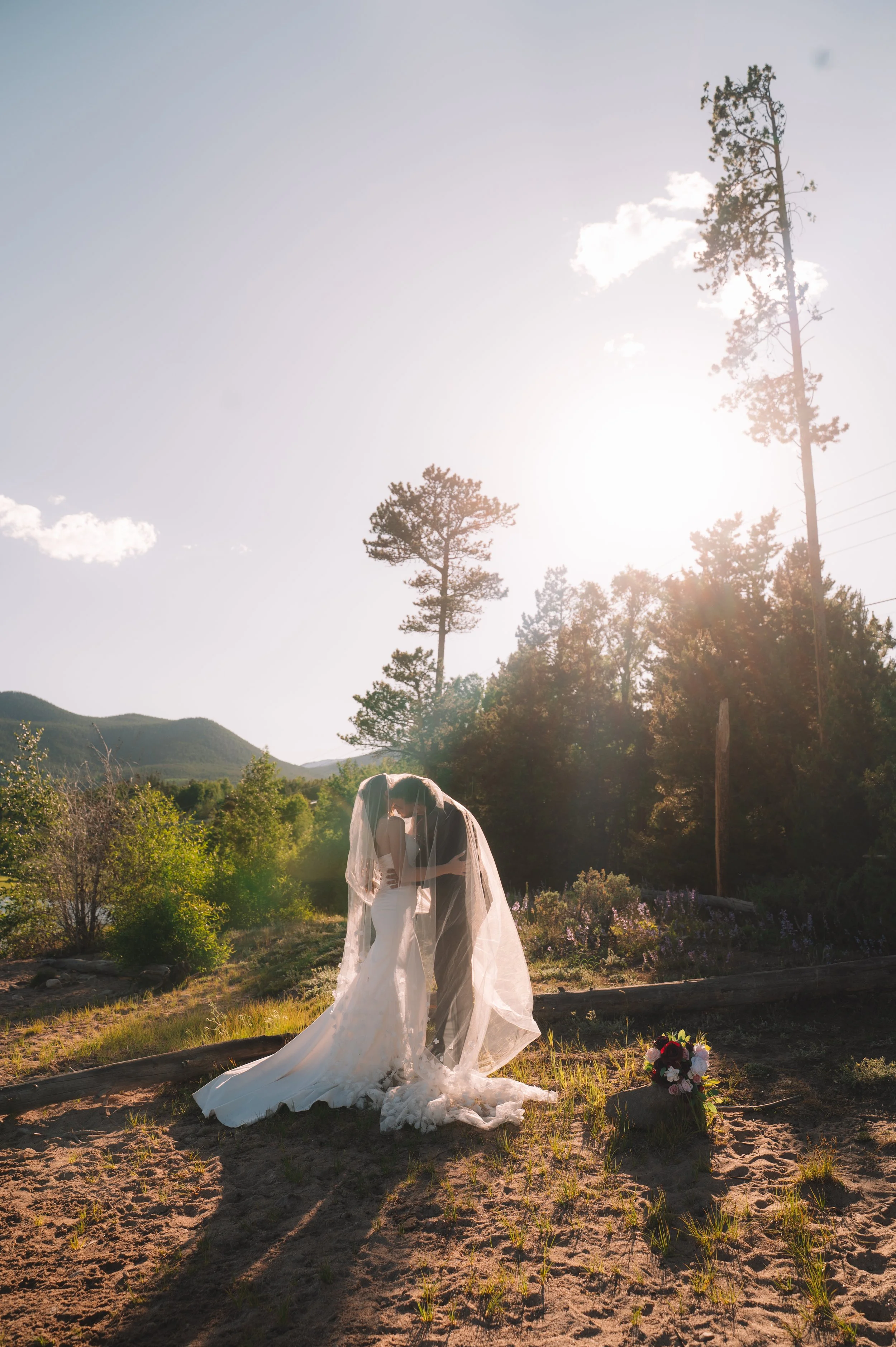 A bride and groom sharing a kiss outdoors during sunset, with a Brecke mountain landscape and tall trees in the background. The bride is wearing a white wedding dress and veil, and the groom is in a gray suit. Flowers are placed on the ground nearby.