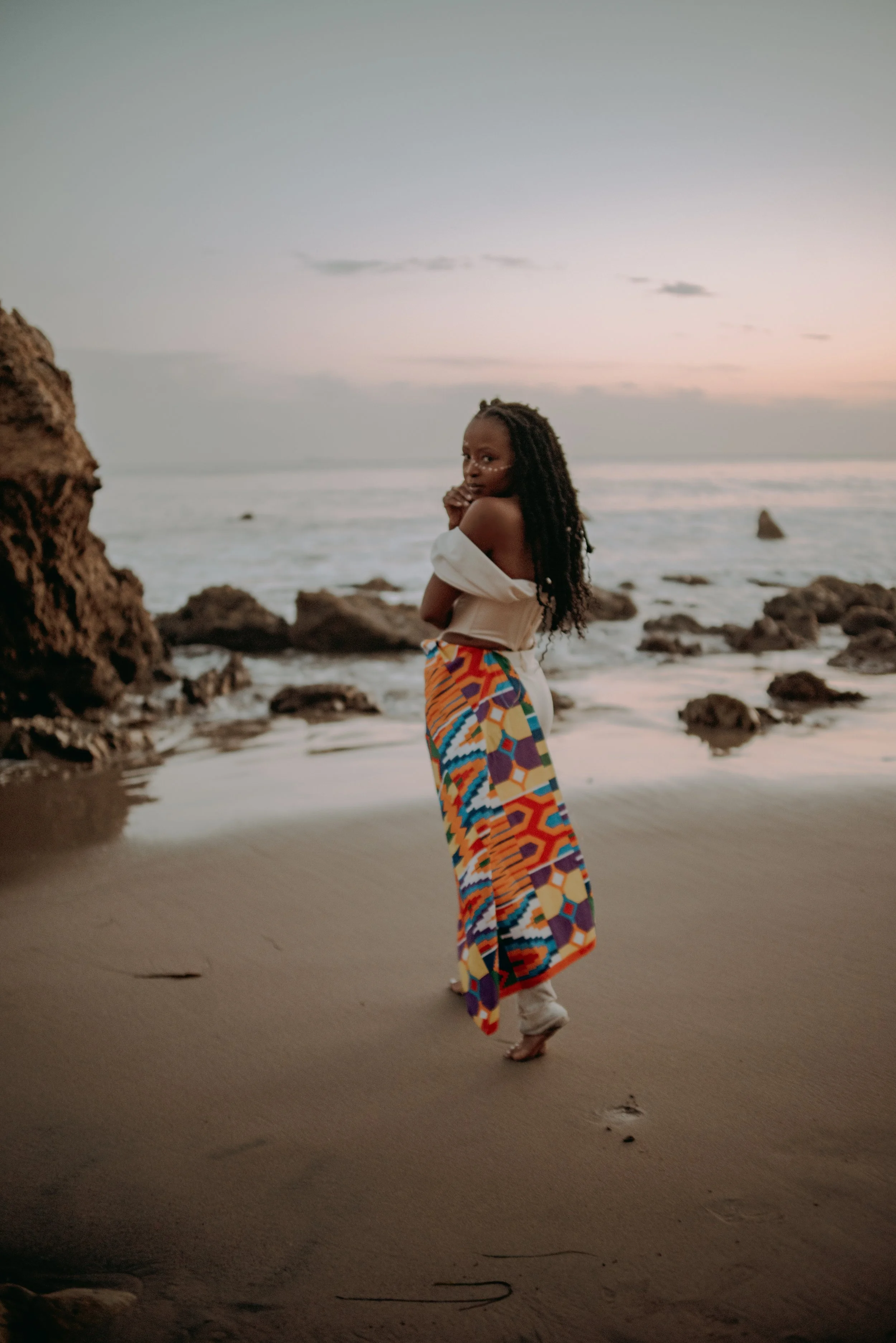 A woman with long braided hair standing on a sandy beach at sunset, wearing colorful patterned pants and a white off-shoulder top, with the ocean and rocks in the background.