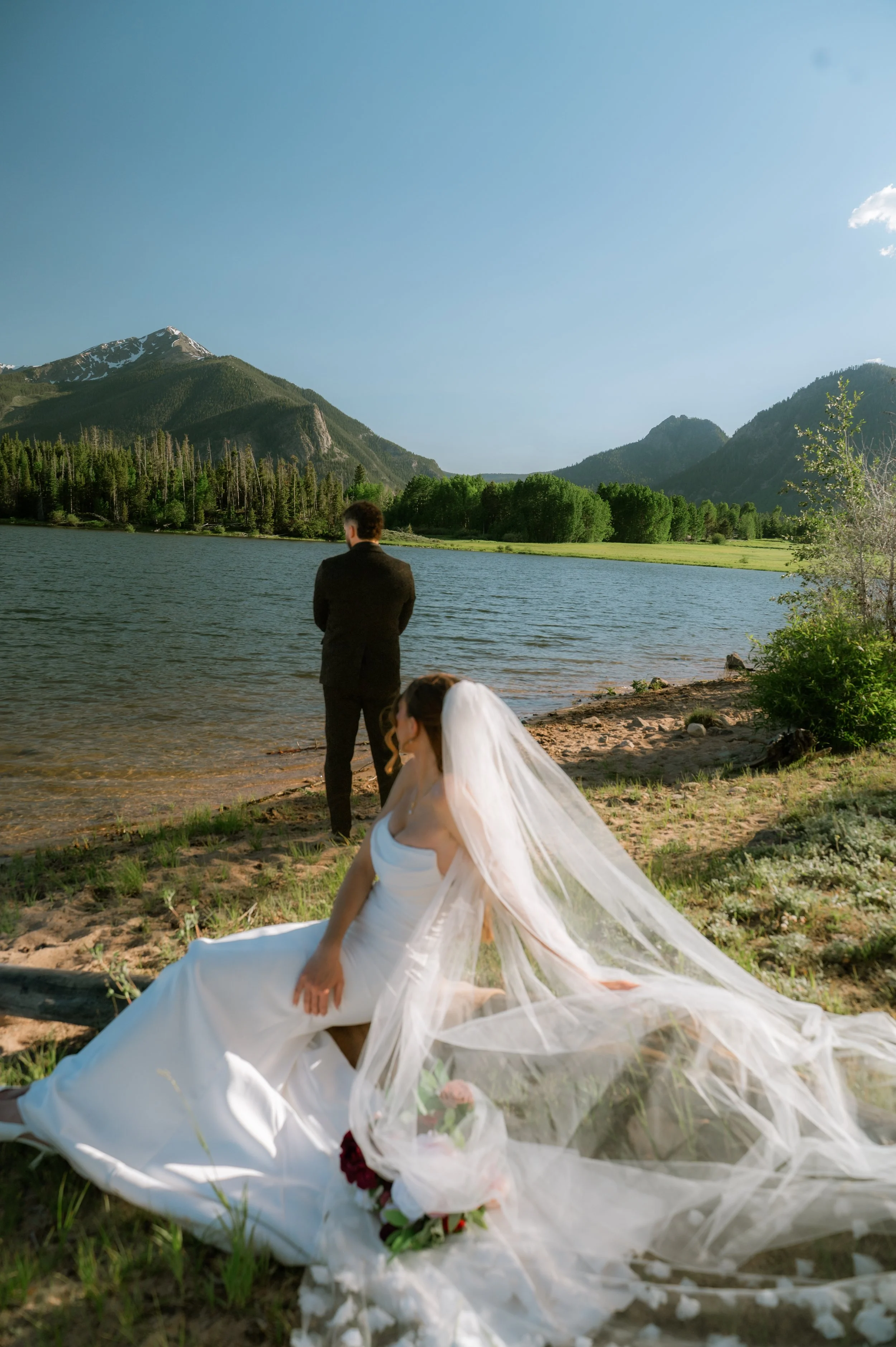 A woman in a wedding dress with a long veil sitting on the ground near a lake, with a man in a dark suit standing nearby, in a scenic mountain landscape with clear blue sky and snow-capped peaks.