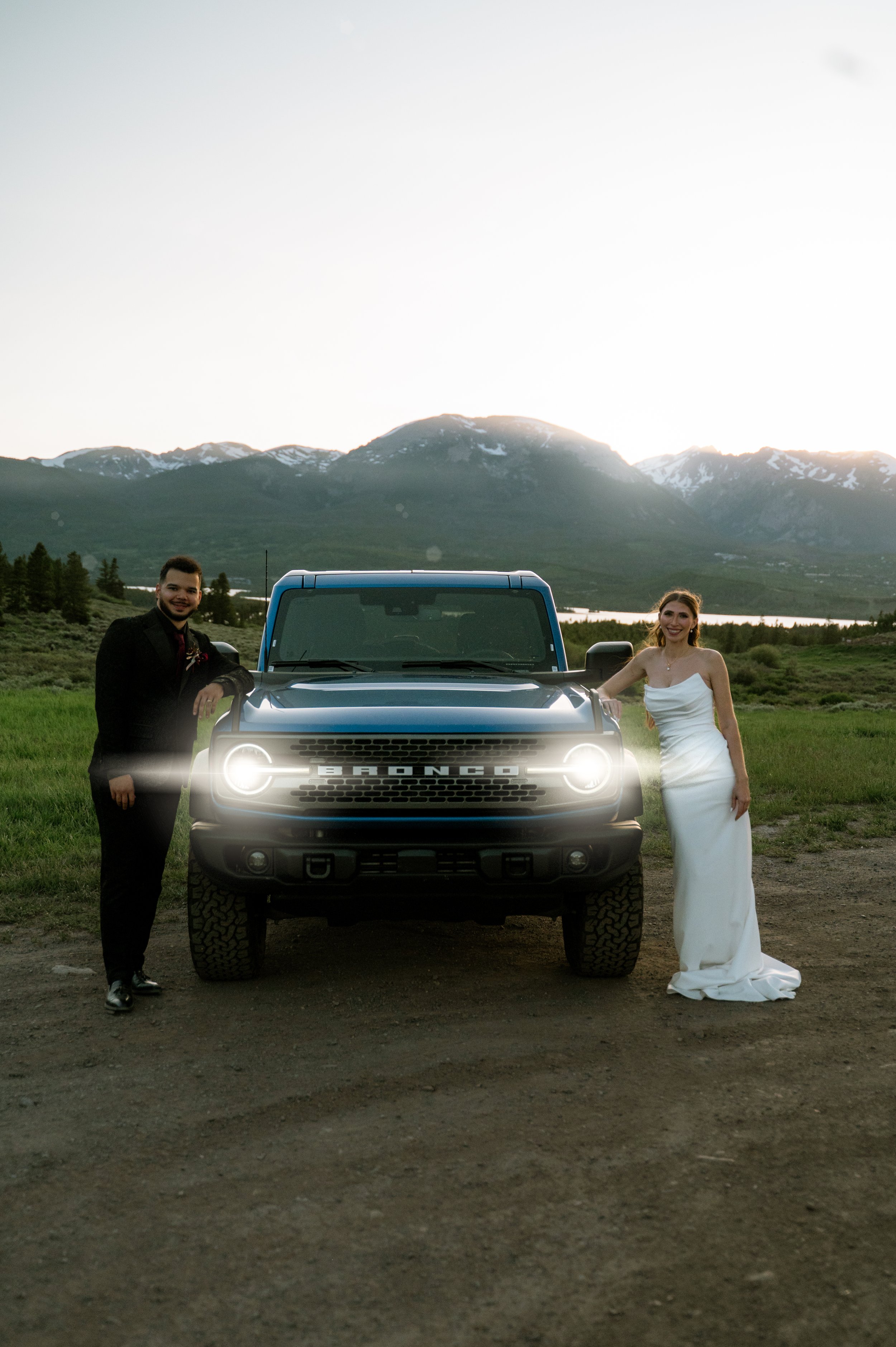 A man in a black suit and a woman in a white wedding dress standing next to a blue Ford Bronco vehicle outdoors with mountains in the background during sunset.
