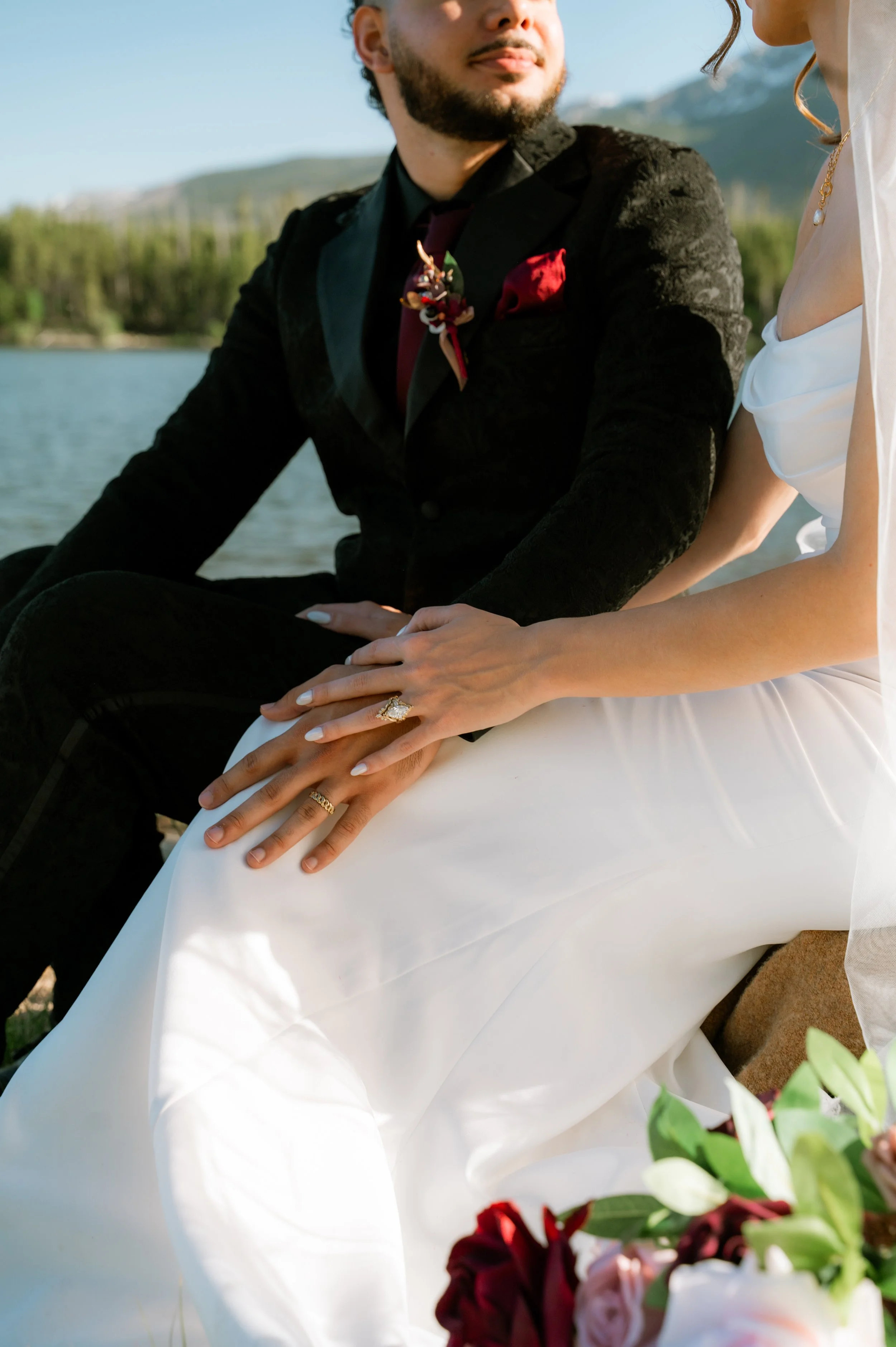 A couple holding hands, sitting outdoors near a body of water with mountains in the background, dressed for a wedding or special occasion.