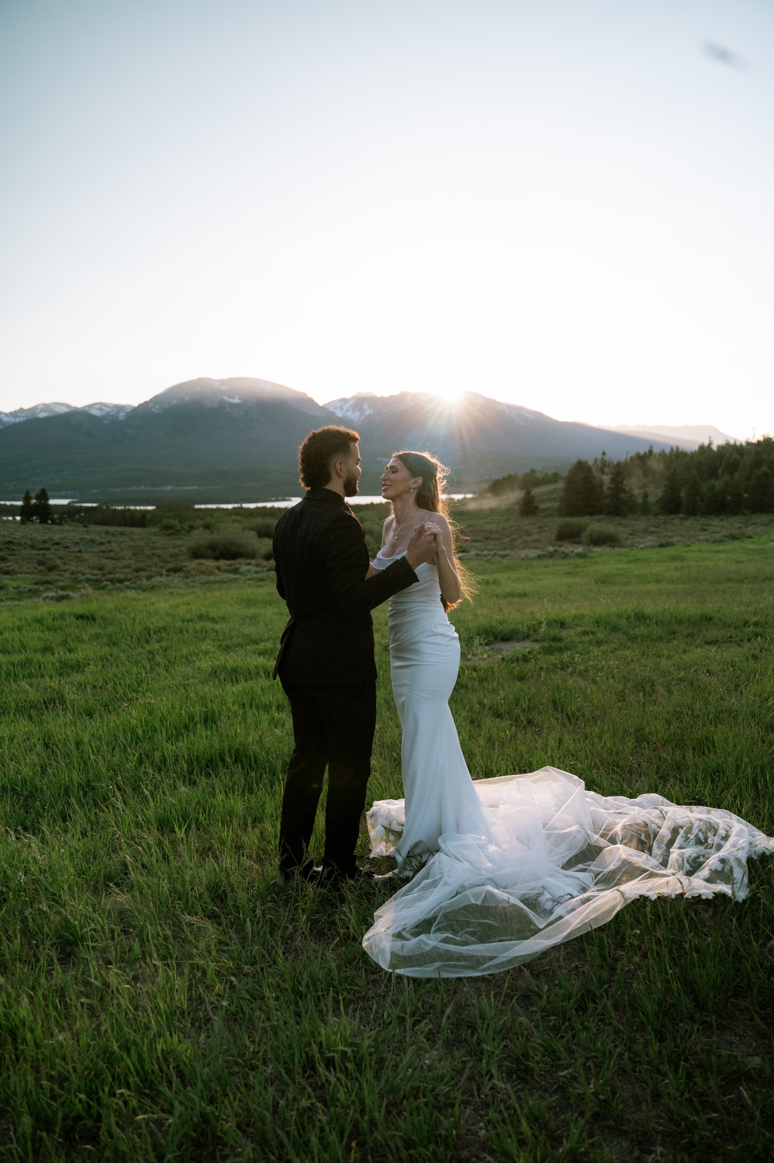 A couple in wedding attire dancing outdoors on green grass with a mountain range and setting sun in the background.