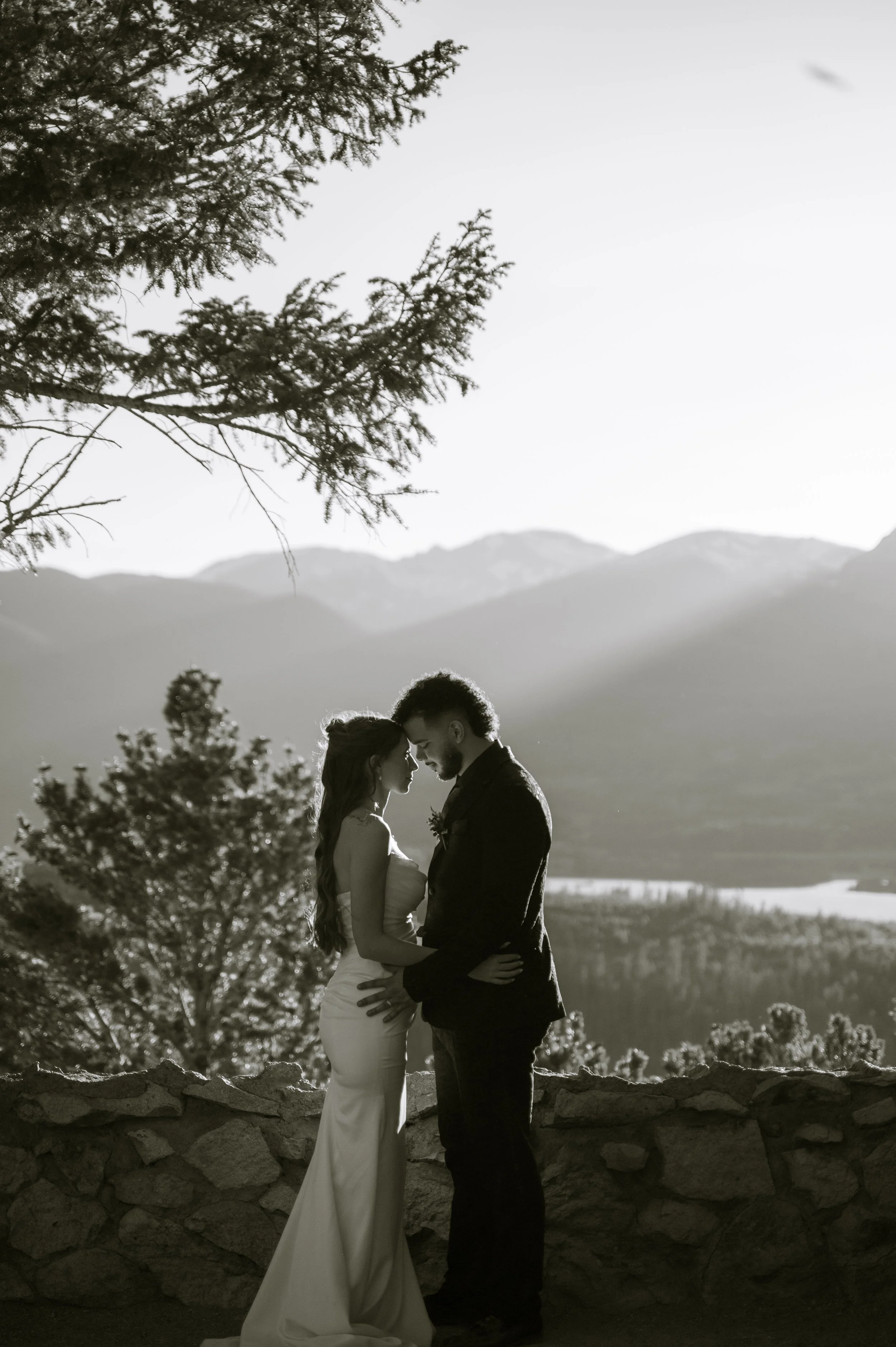 A black-and-white photo of a couple standing close together outdoors with aspen mountains and trees in the background, their faces touching, likely during a romantic moment.