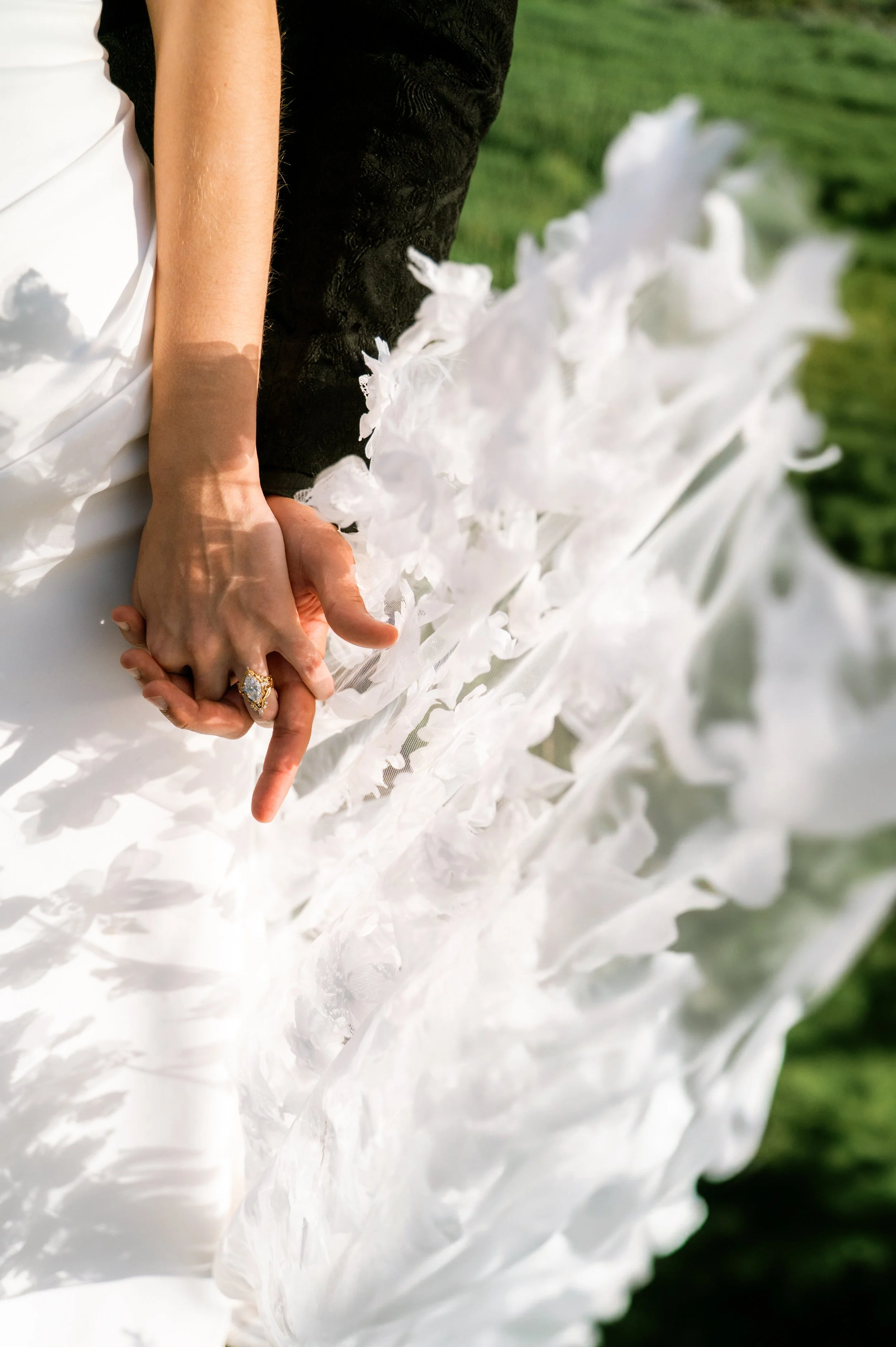 Close-up of two people holding hands, one wearing a white wedding dress with a ring on the finger, and the other dressed in a black suit, with a white floral decoration in the background, outdoors on grass.