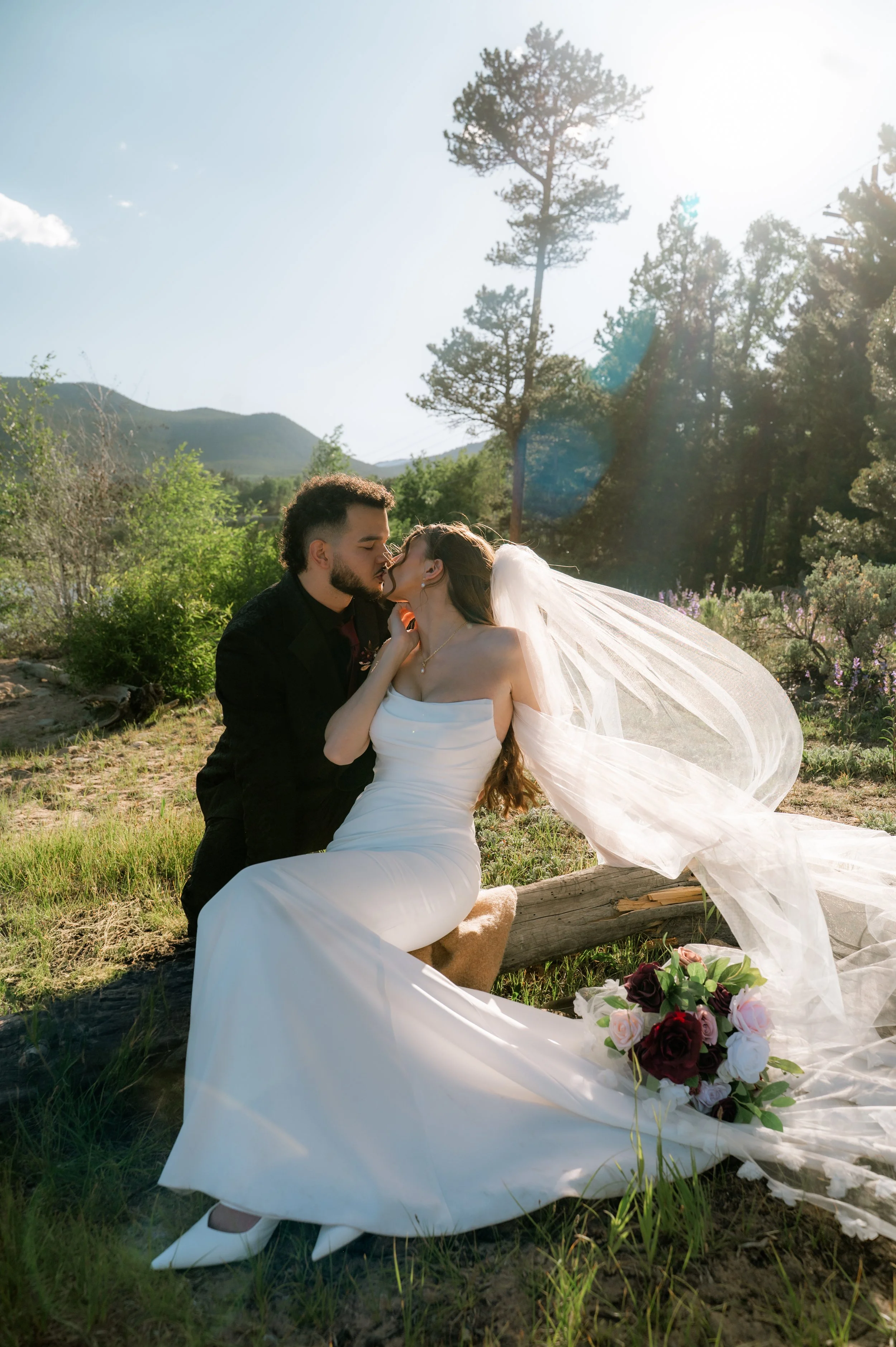 A couple dressed in wedding attire, the man in a black suit and the woman in a white wedding dress, sharing a kiss outdoors in a natural setting with trees, mountains, and the sunlight shining behind them.