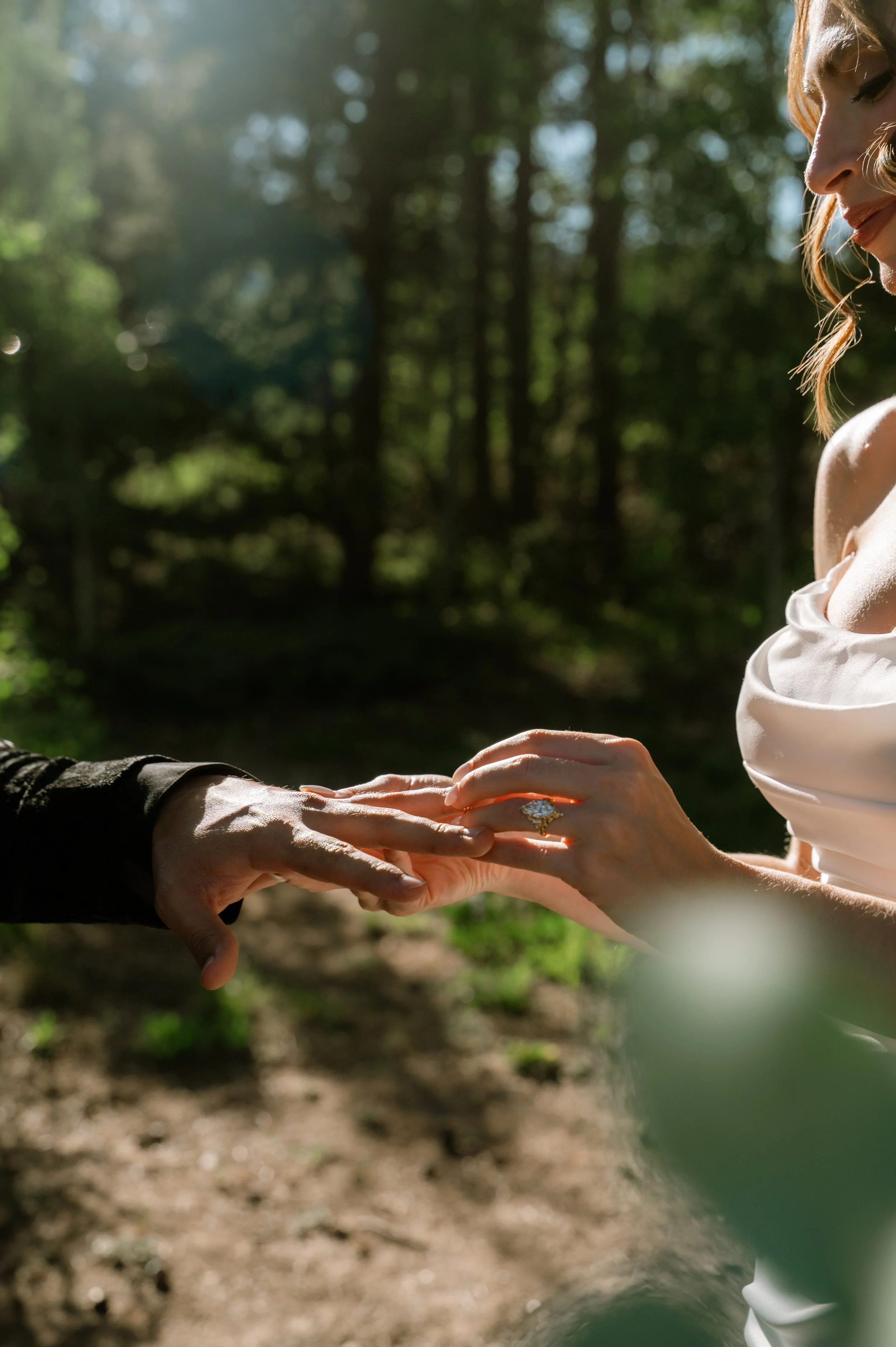 A couple exchanging wedding rings outdoors in a wooded area with sunlight filtering through the trees.