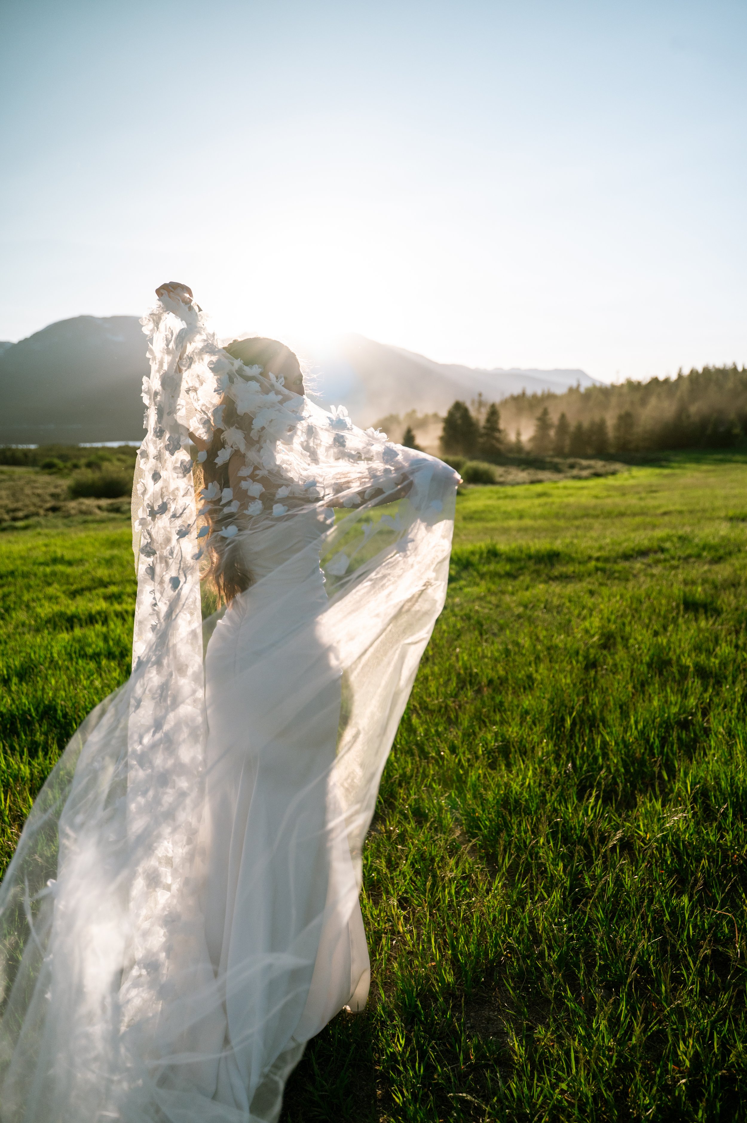 A woman in a white dress and a transparent veil standing in a green field with mountains in the background, illuminated by the setting sun.