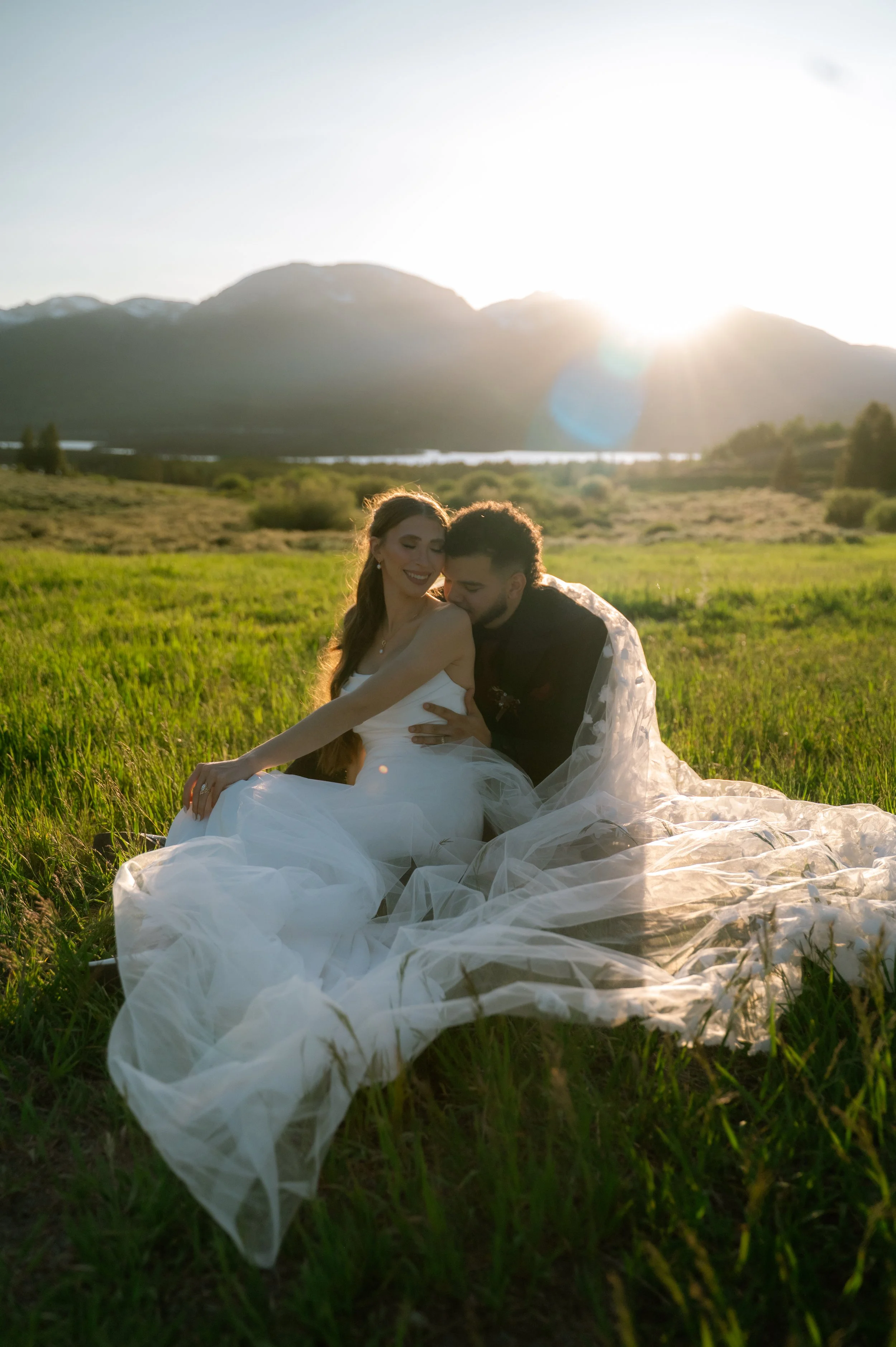 A bride and groom sitting close together in a green field with mountains in the background and the sun setting behind them.
