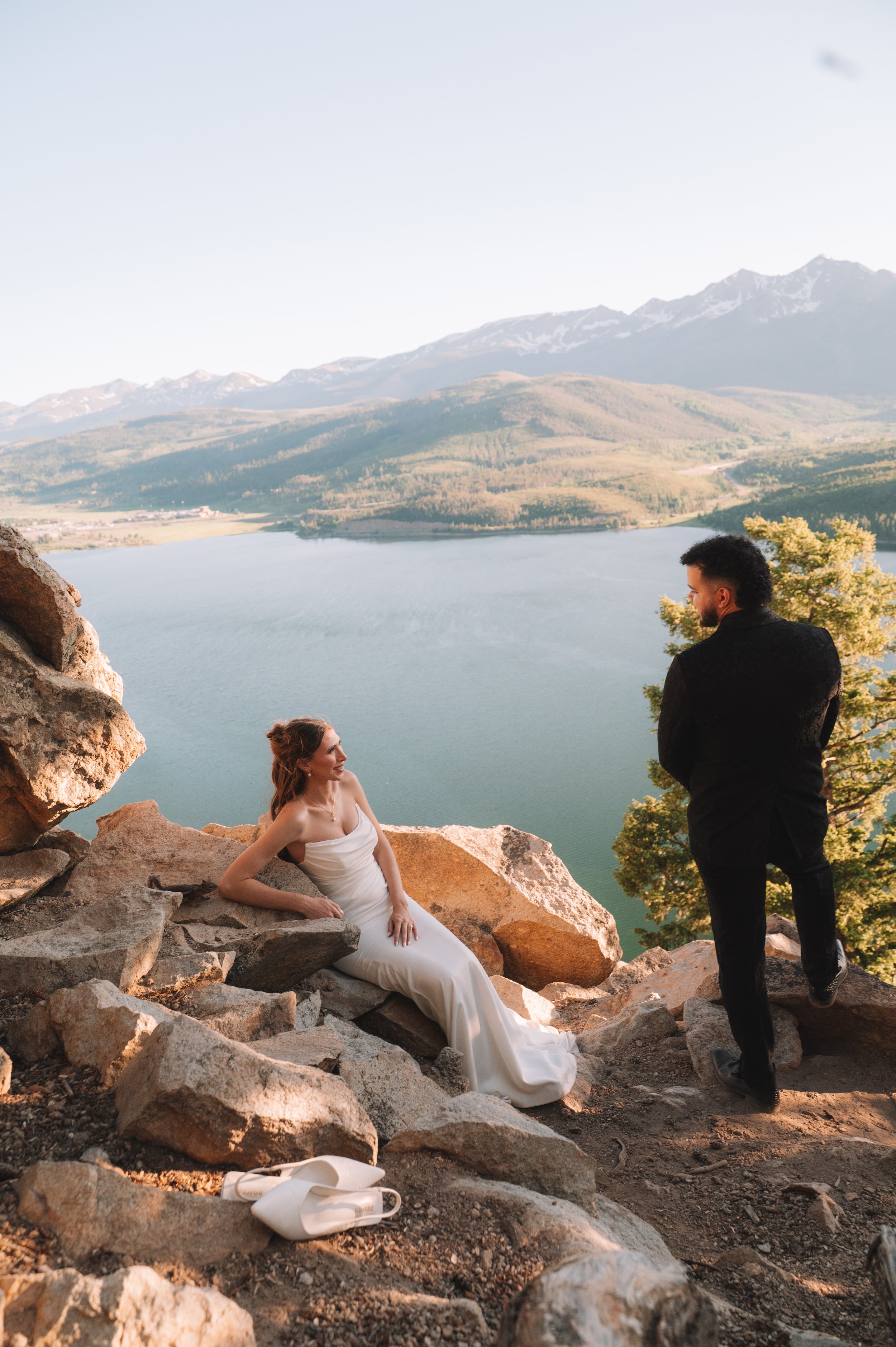 A woman in a white dress sitting on rocks by a lake with mountains in the background, and a man in black standing nearby. The woman is smiling and looking at the man.