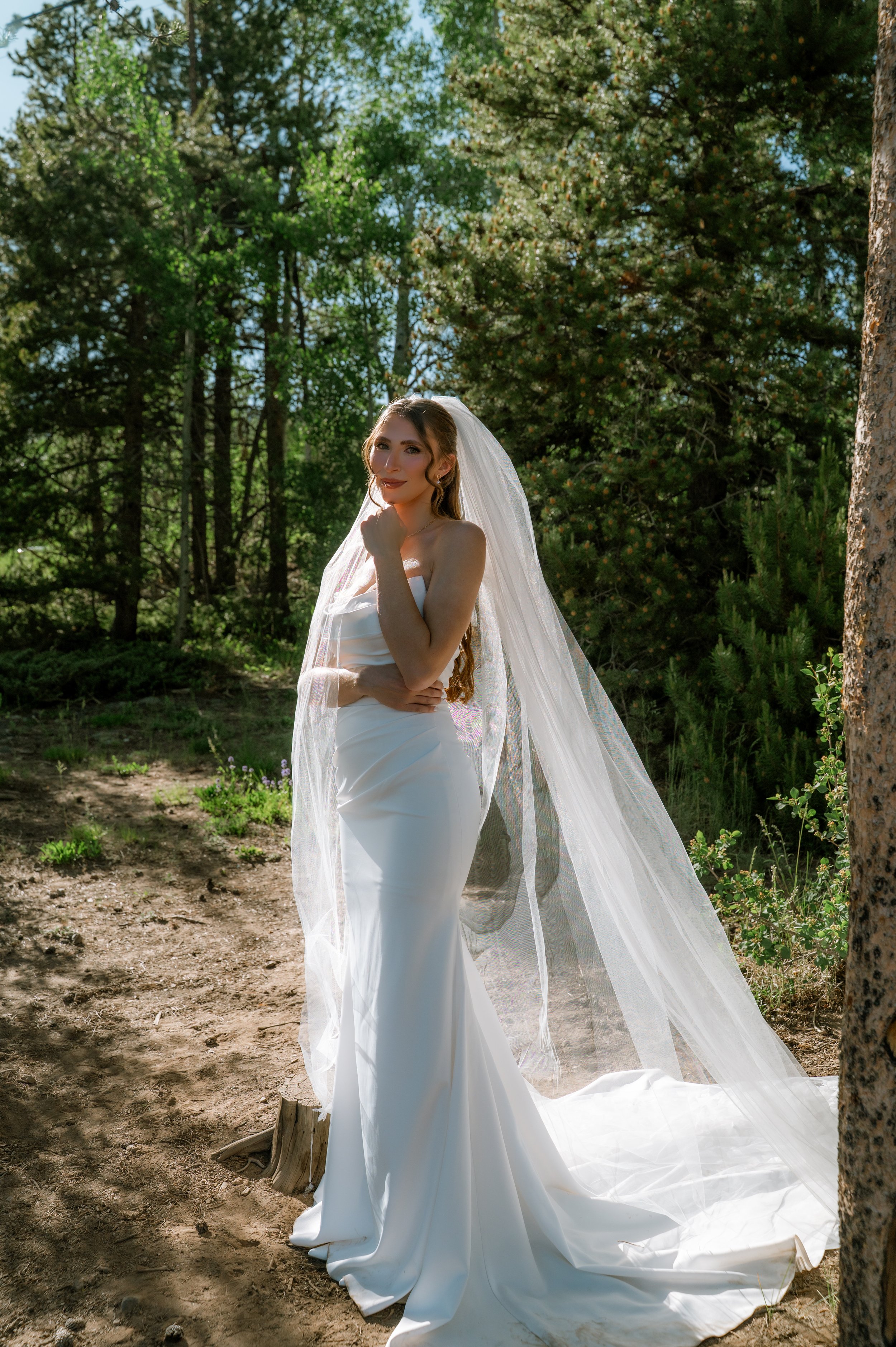 A high fashion style bride in a white wedding dress and veil stands outdoors among trees in mountains, with sunlight shining on her.