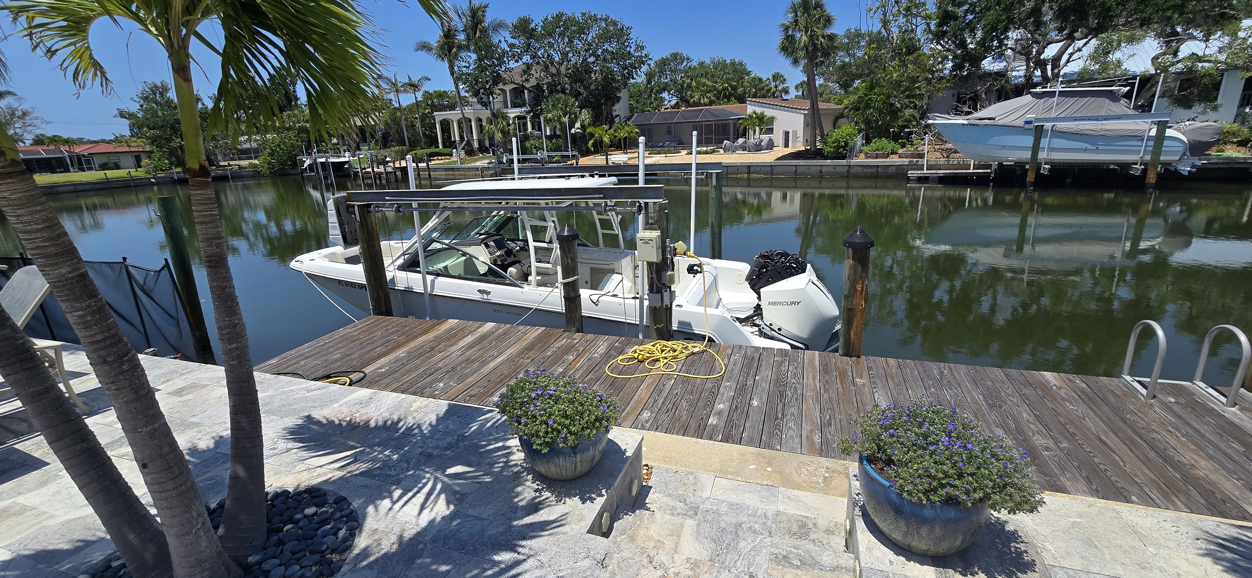 A dock with a boat tied up and a scenic waterway with homes and boats in the background, under a clear blue sky.