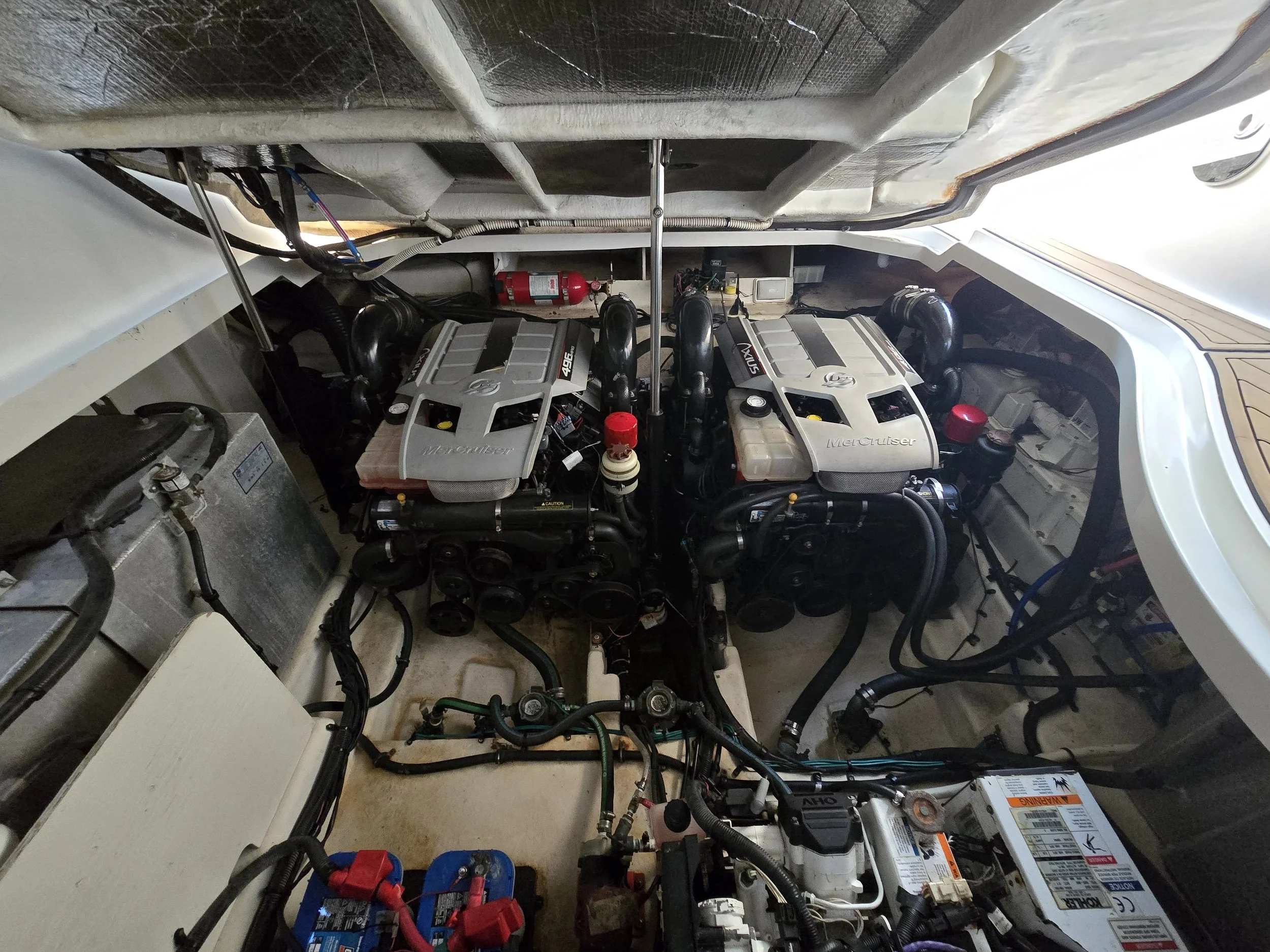 An engine compartment with two Mercuriser engines, a battery, a fire extinguisher, and various hoses and components.