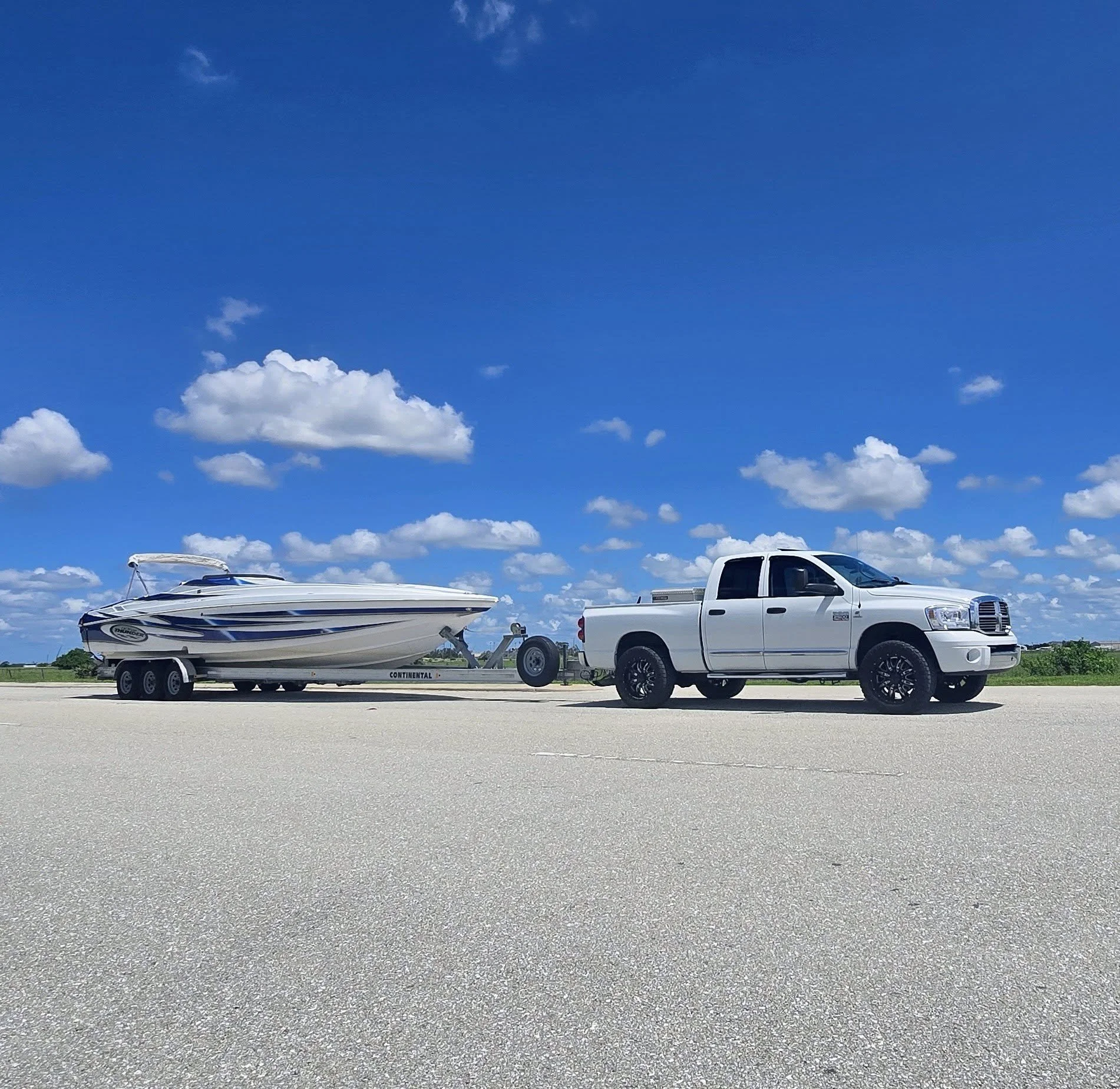 A white pickup truck towing a boat on a trailer, parked on an open paved area under a blue sky with scattered clouds.