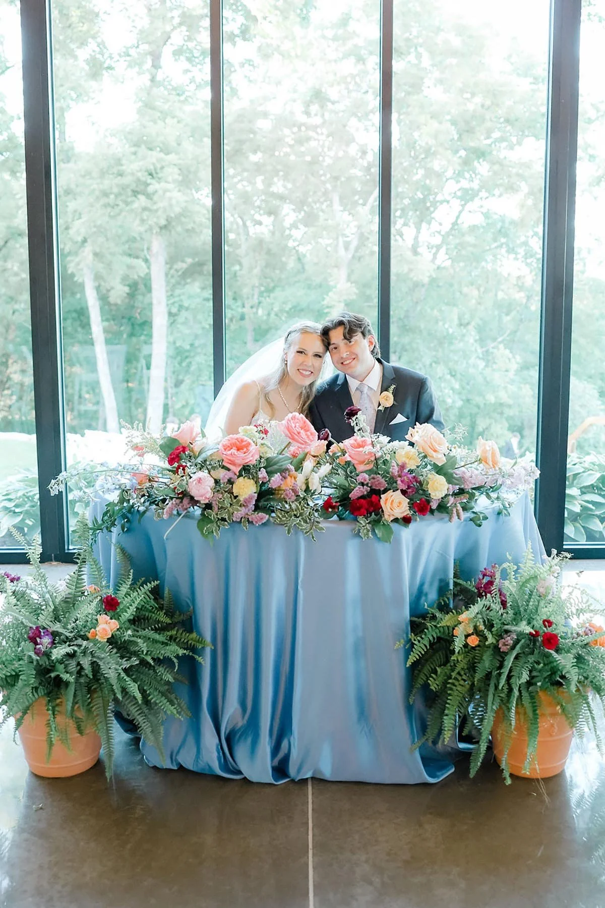  Newlyweds and sitting at their sweetheart table with blue linens, and lots of colorful florals on top. 