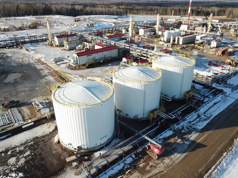An industrial facility with three large, white cylindrical storage tanks connected by pipes. Snow covers the ground and tanks. There are various buildings, equipment, and vehicles around the site, with a forest and snow-covered landscape in the background.
