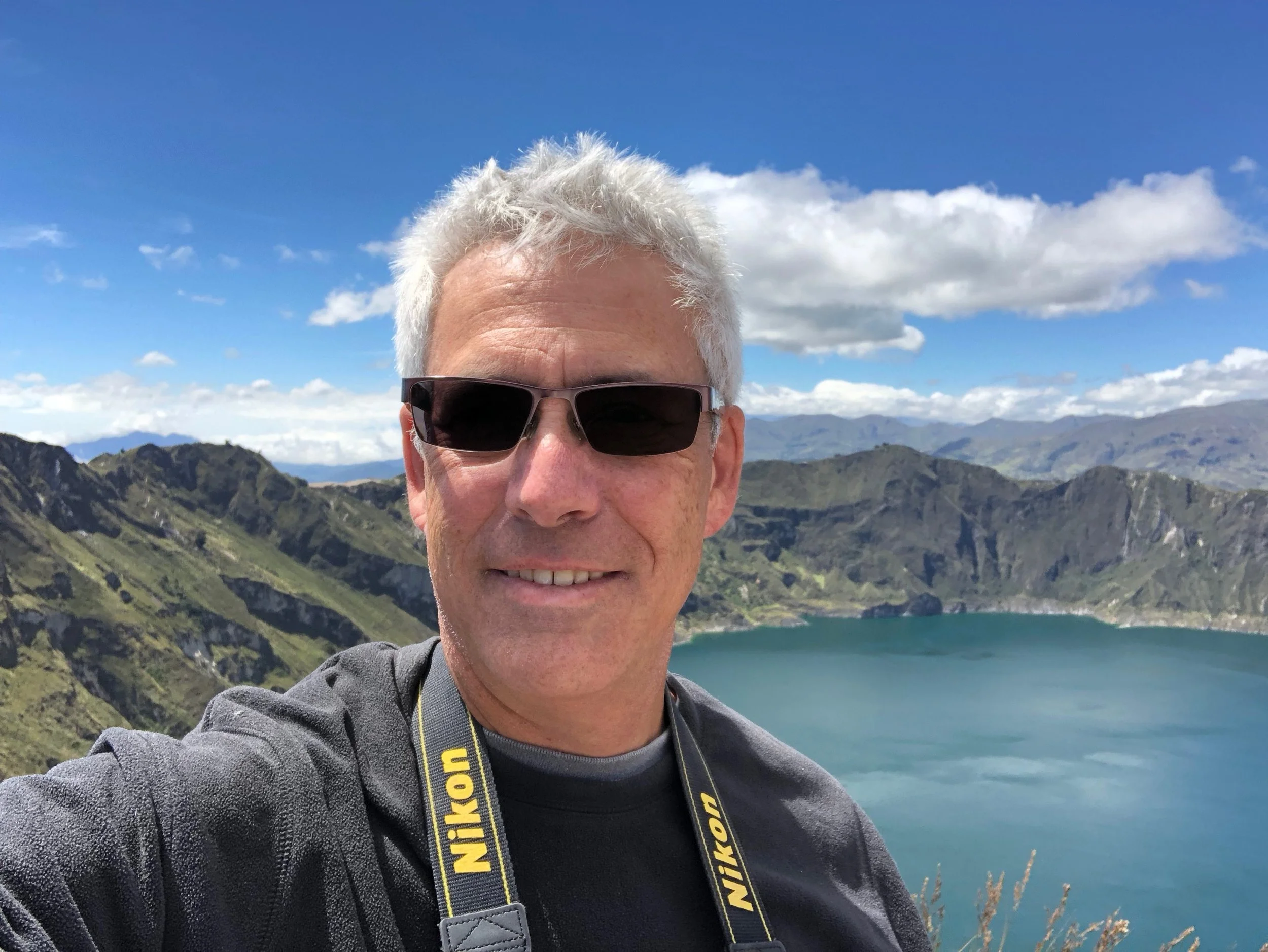 Hombre adulto de pelo blanco y con gafas de sol, posa sonriente delante de un lago. De su cuello cuelga una camara de fotos y en el fondo vemos montañas y un cielo azul con algunas nubes.