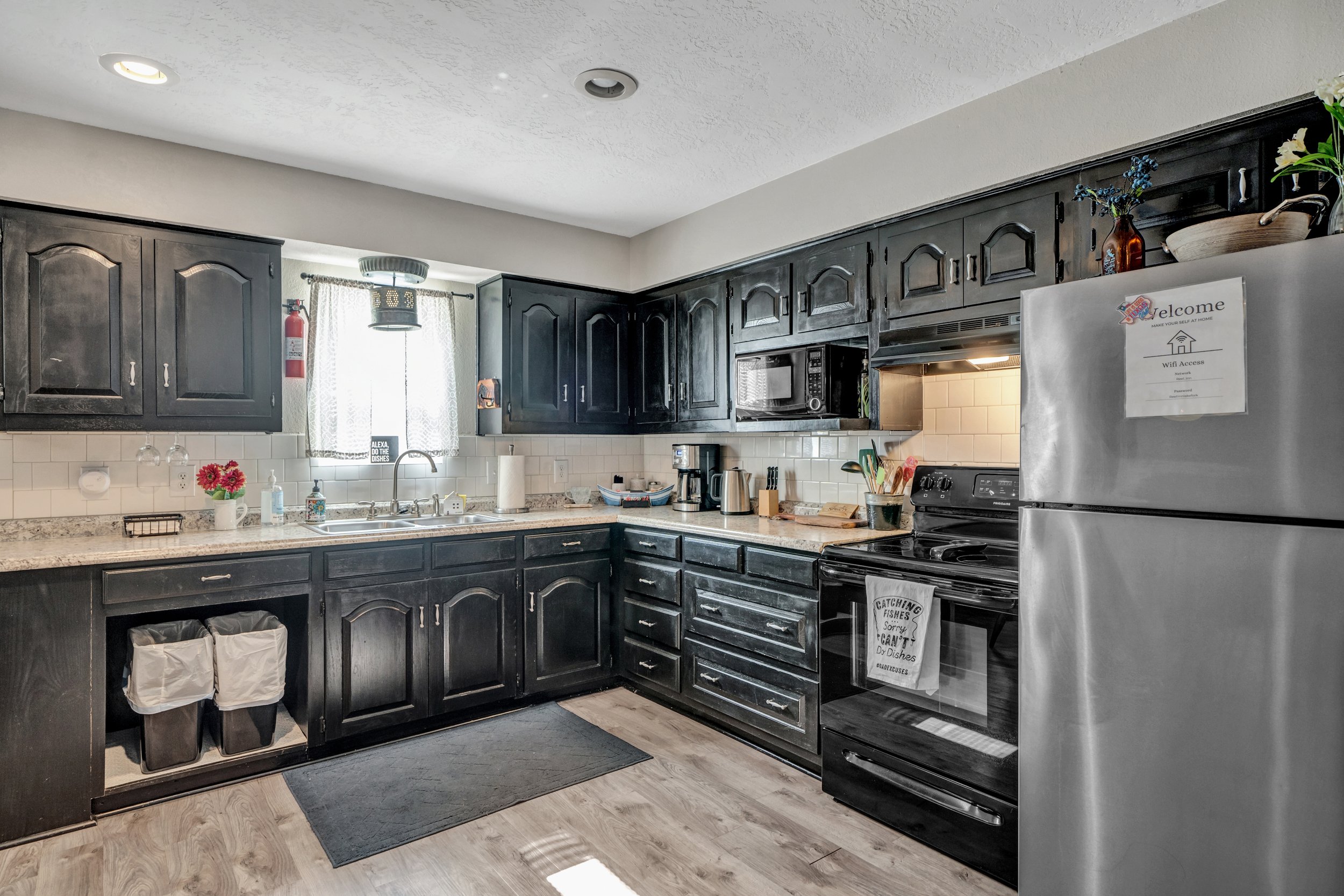 Modern kitchen with dark cabinets, stainless steel appliances, and a tiled backsplash. Features a sink under a window, microwave, oven, and refrigerator. Countertops display small appliances and decor. Light wood flooring.