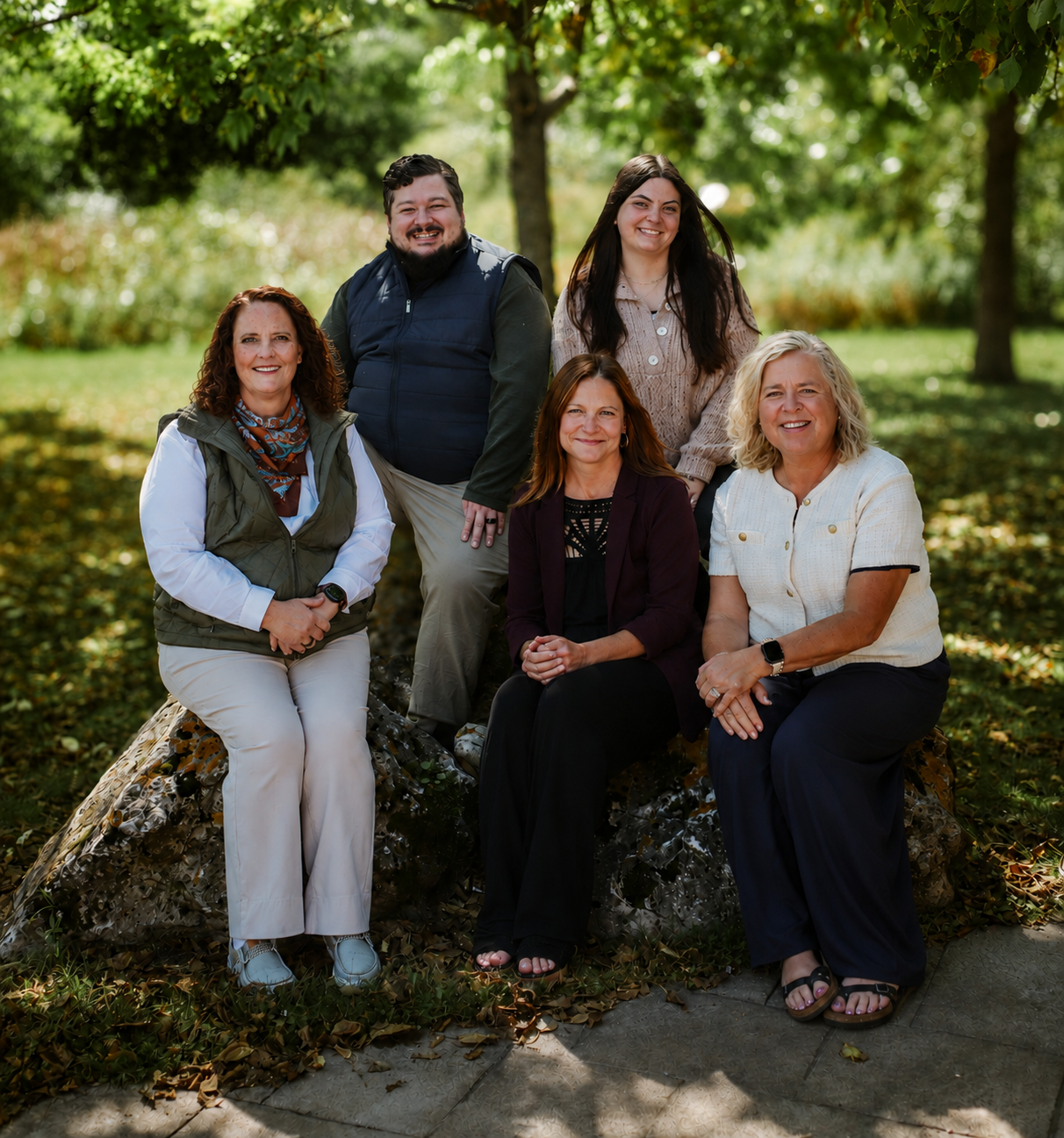 Group of six people outdoors, sitting and standing on and around a large rock, surrounded by trees with green foliage, in a park on a sunny day.
