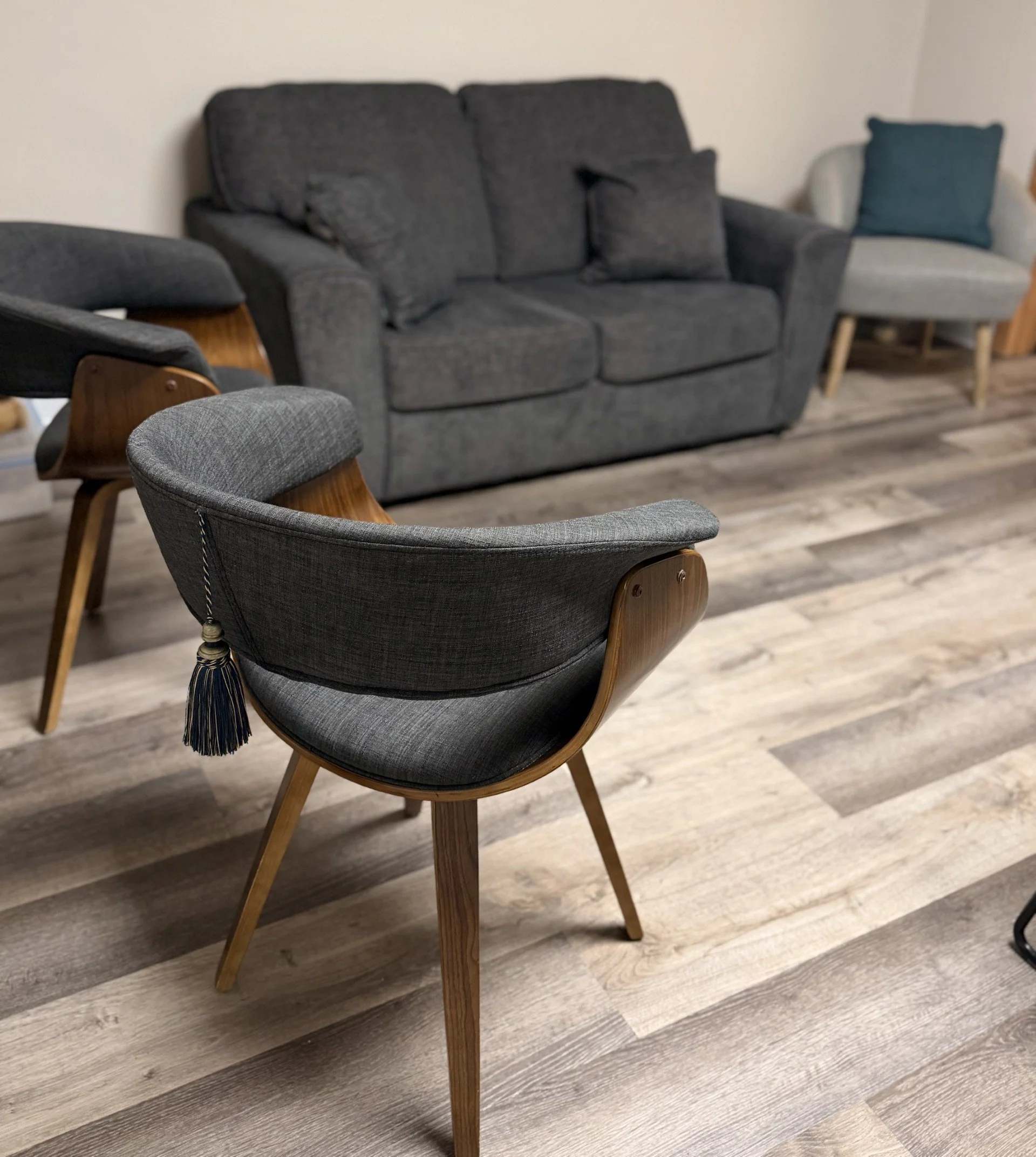 A modern living room with a gray upholstered armchair in the foreground and a dark gray fabric sofa with pillows in the background, along with a white armchair with a blue pillow, and wood flooring.