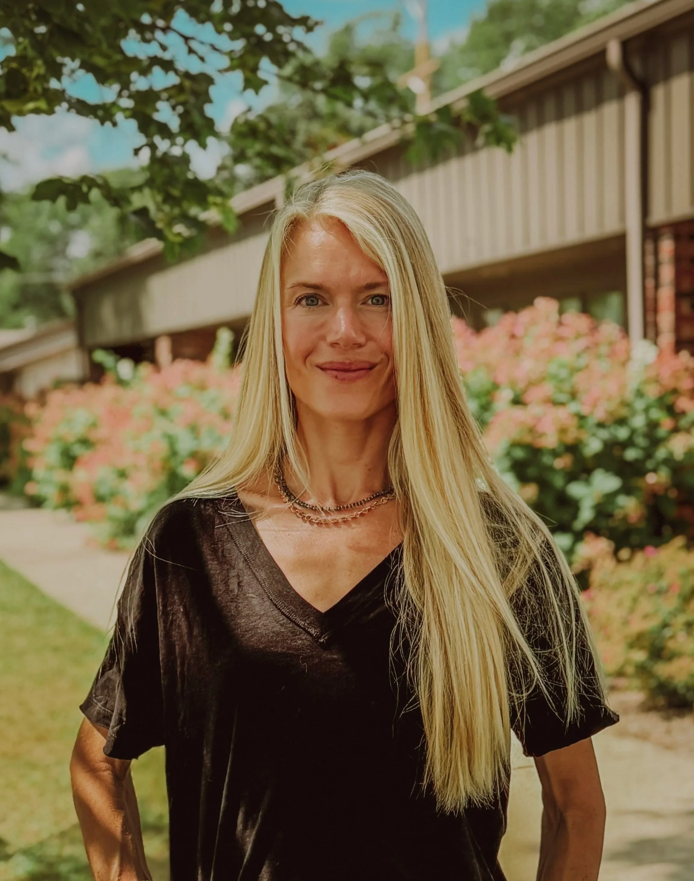A woman with long blonde hair standing outdoors in front of a hedge with pink flowers, a wooden building, and trees, smiling at the camera.