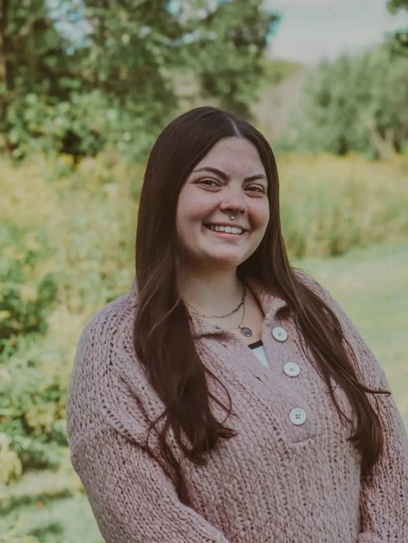 A young woman with long dark hair, wearing a beige knit zip-up sweater, standing outdoors in a park with green grass and trees in the background.