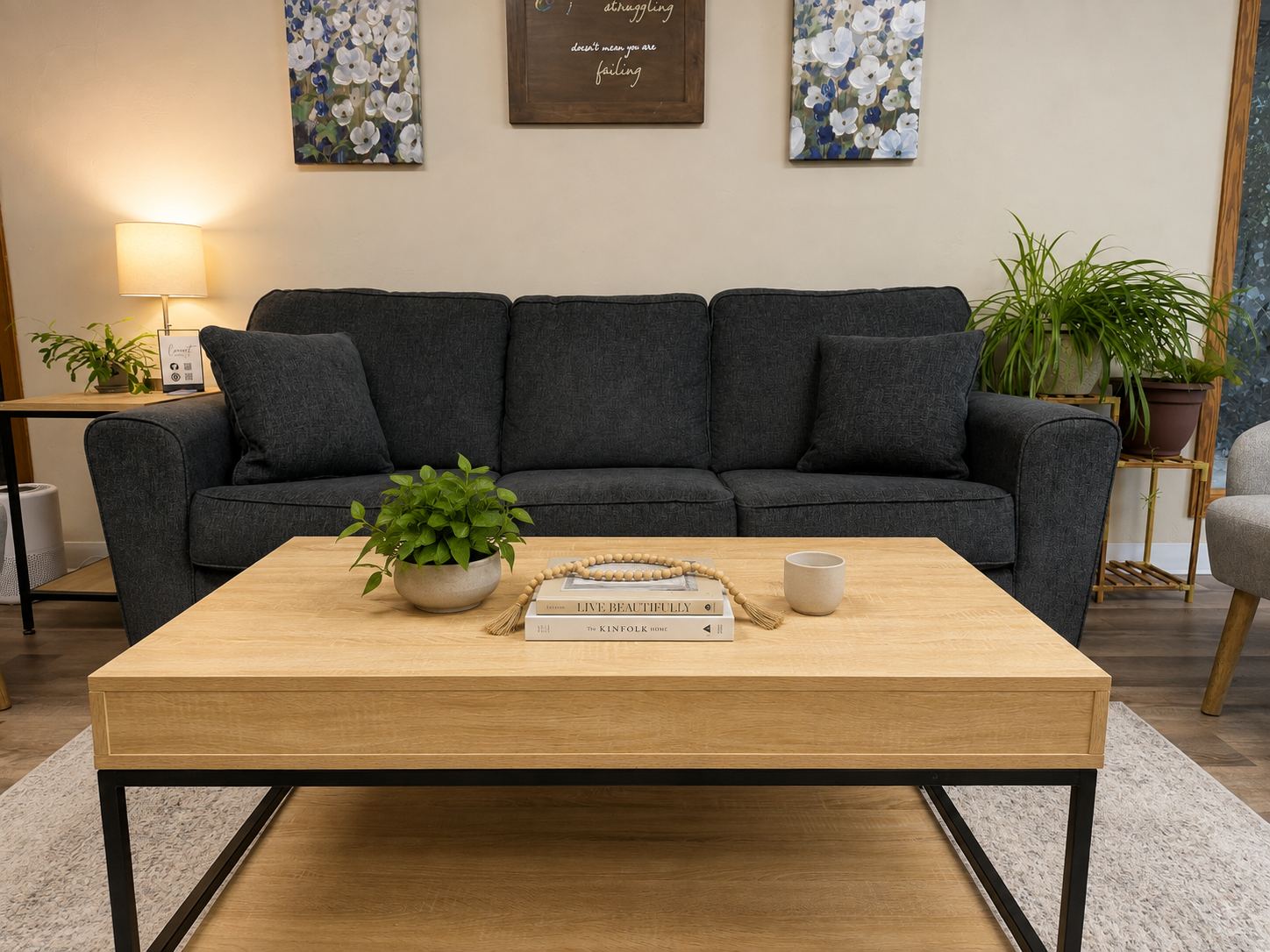 A living room with a dark gray sofa, a light wooden coffee table with books and decorative items, potted plants, a table lamp, and framed artwork on the wall.