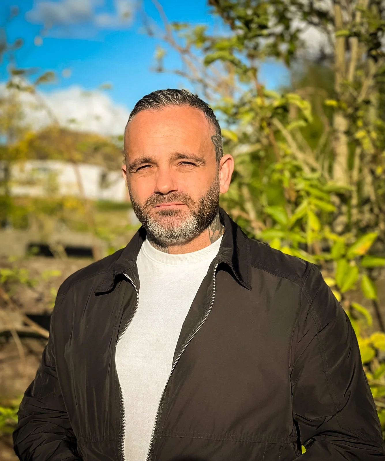 Bearded tattooed man in a black jacket standing outdoors in warm sunlight