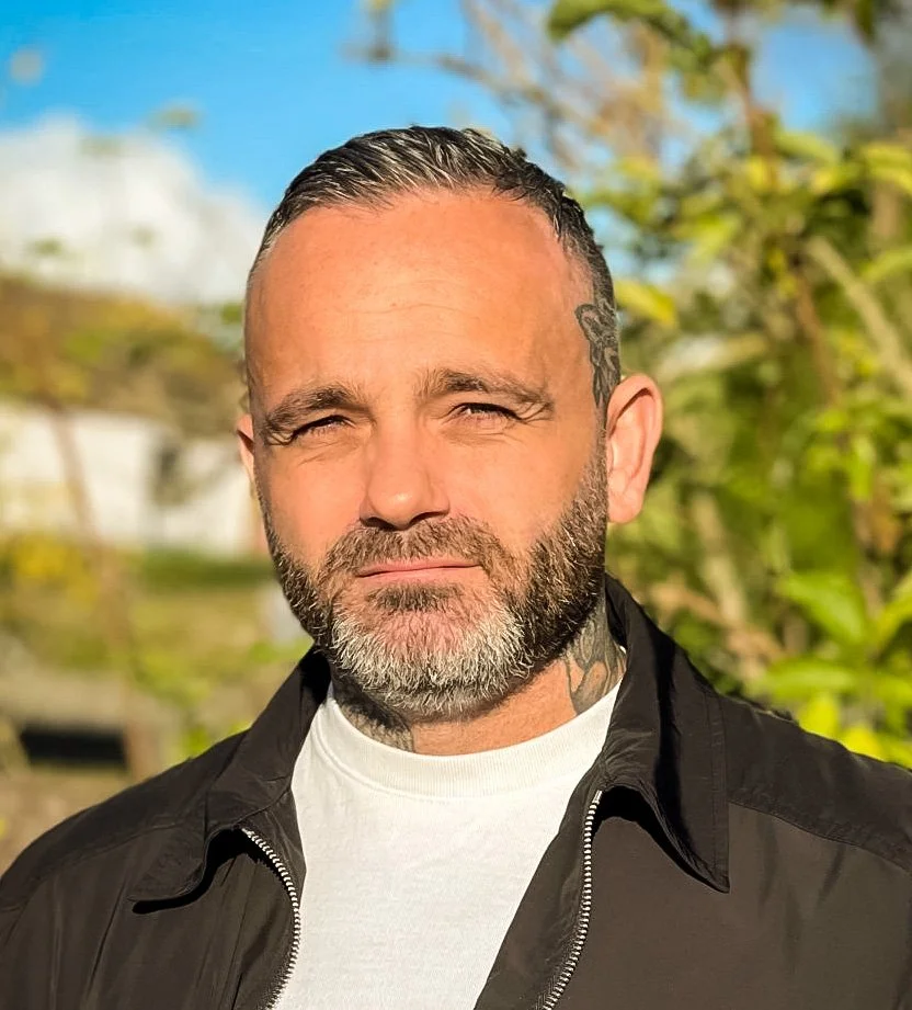 Portrait of a bearded man with neck tattoos wearing a black jacket outdoors in sunlight