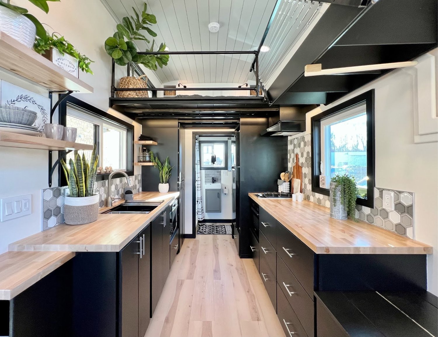 Interior of a modern, tiny home kitchen with black cabinets, light wood countertops, hexagonal backsplash tiles, and potted plants. Features open shelving, natural light, and a loft with railings above.