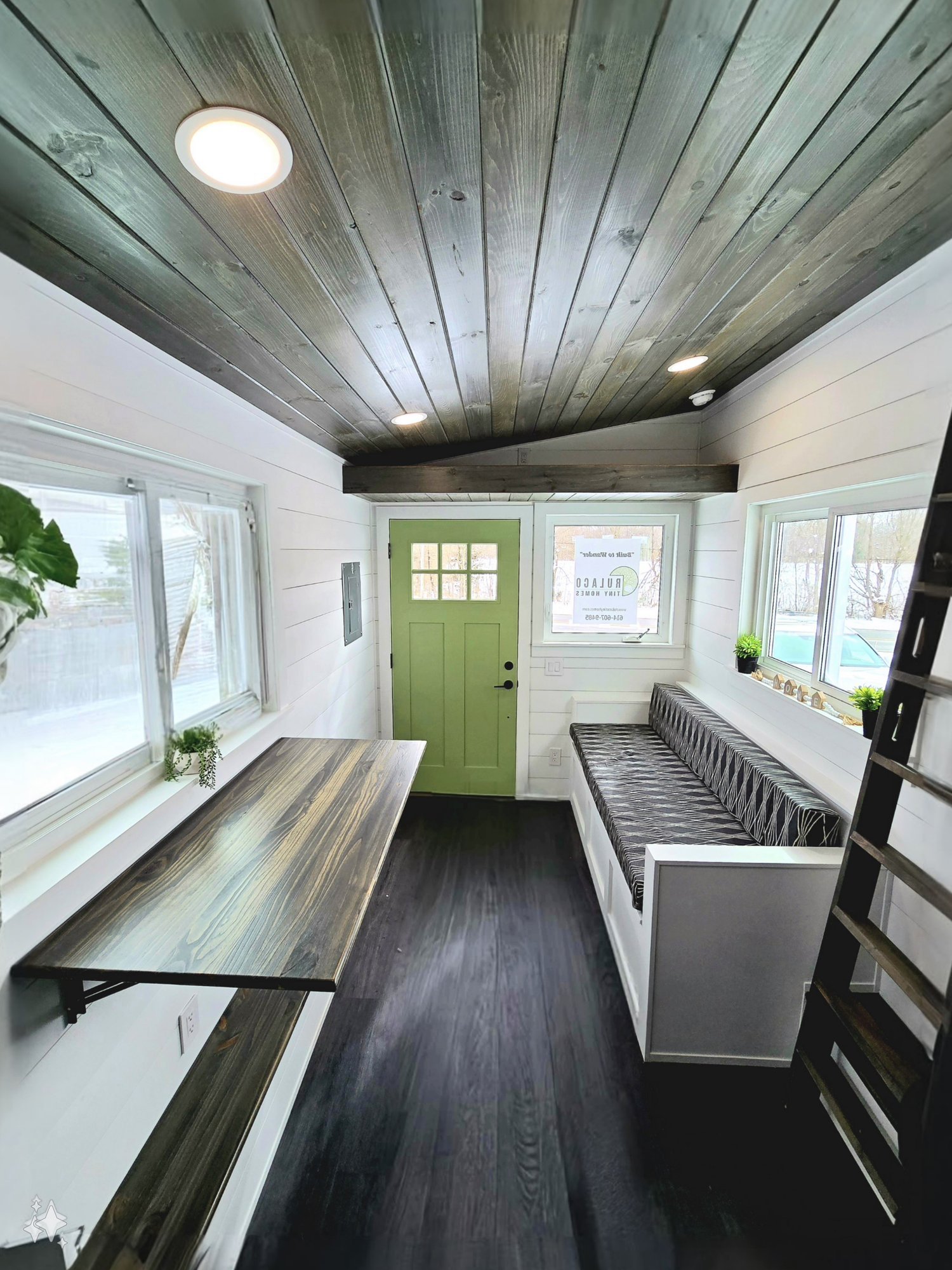 Interior of a small modern room with a wood-paneled ceiling, dark floor, green door, bench seating, and folding table by a window. Minimalist decor with small plants on ledges.