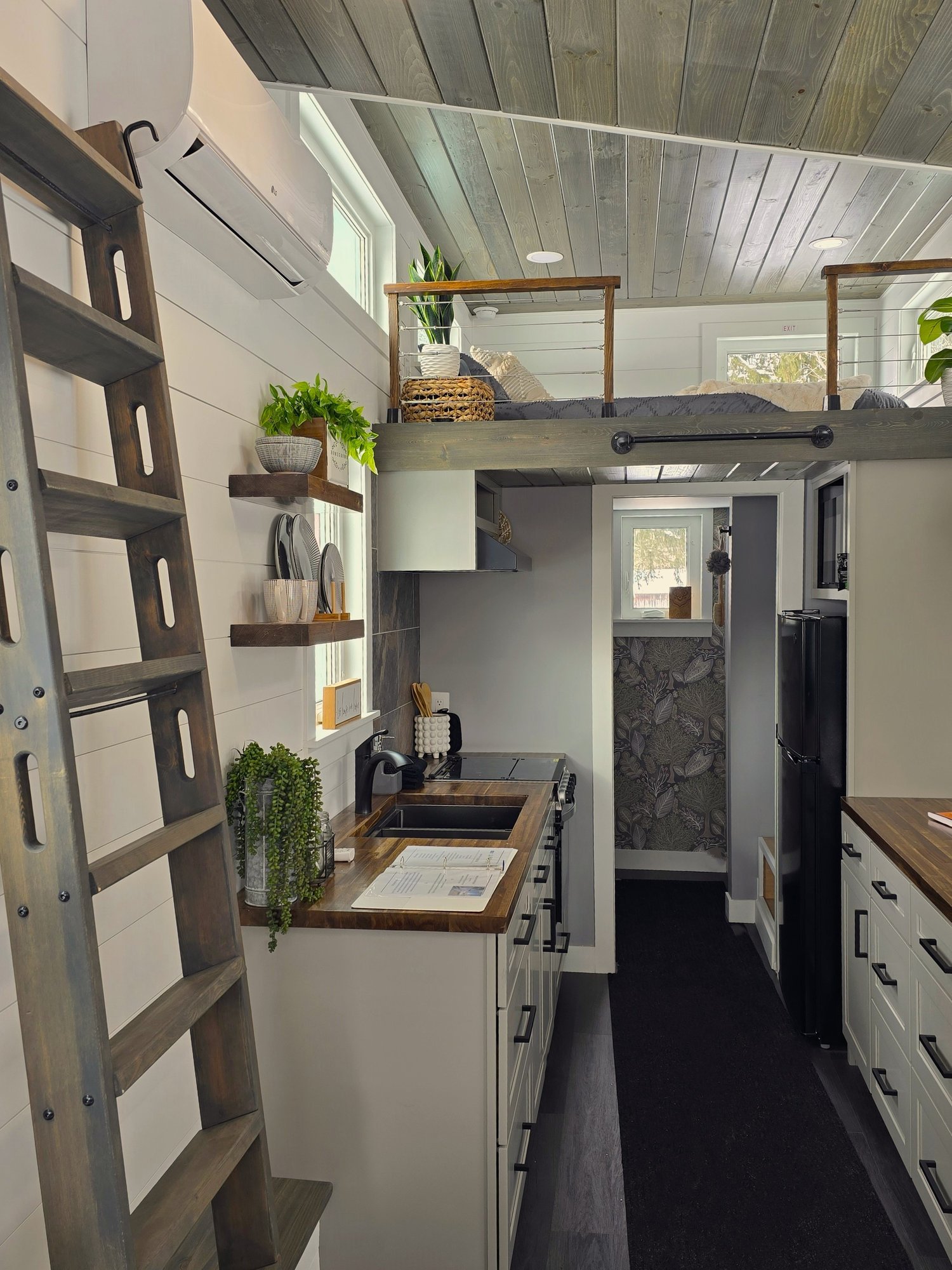 Interior of a tiny house with a modern kitchen, wooden countertops, shelves, and a ladder leading to a loft with plants and bedding.