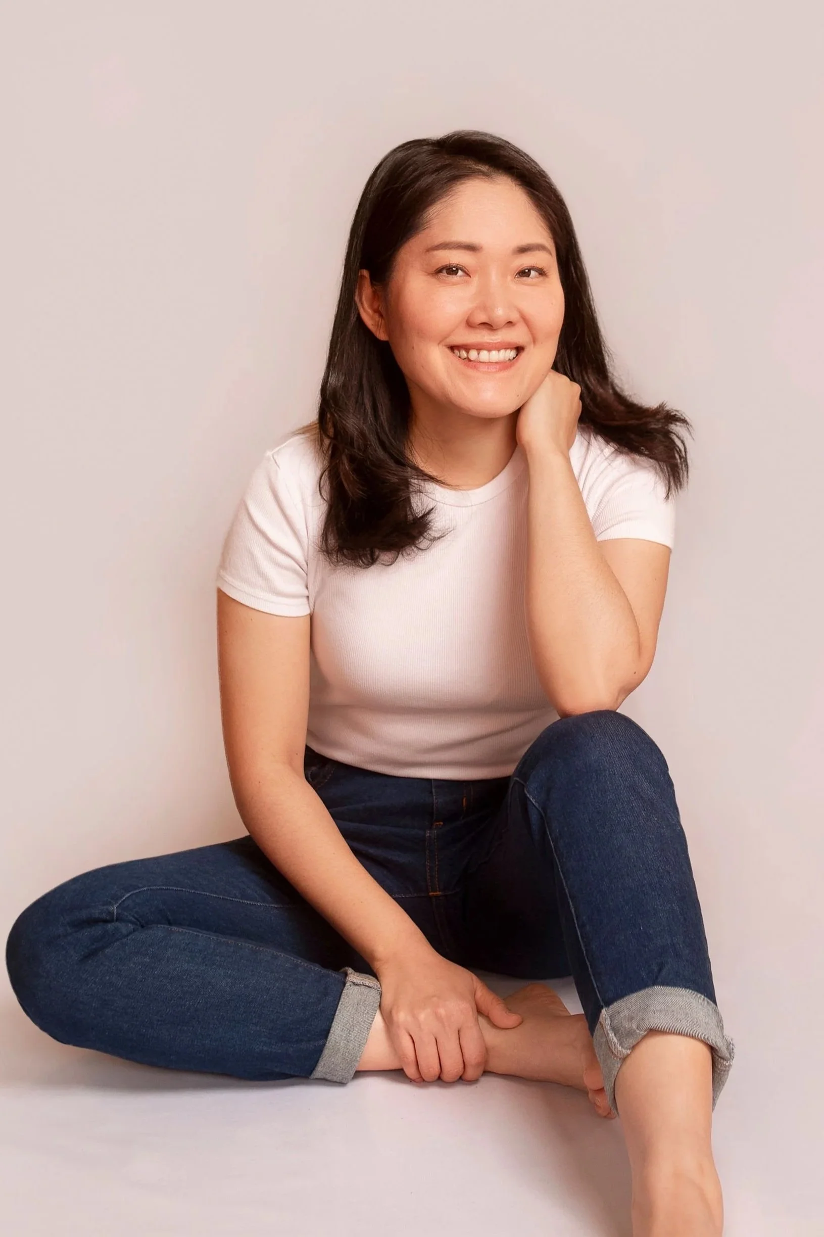 A woman with shoulder-length dark hair, wearing a white t-shirt and dark jeans, sitting on the floor against a plain light-colored wall, smiling at the camera with her left hand resting on her neck and her right hand on her ankle.