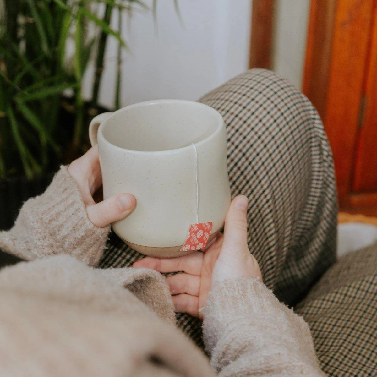 Person holding a ceramic mug while sitting with legs crossed, dressed in plaid pants and a beige sweater, with green plants and wooden furniture in the background.